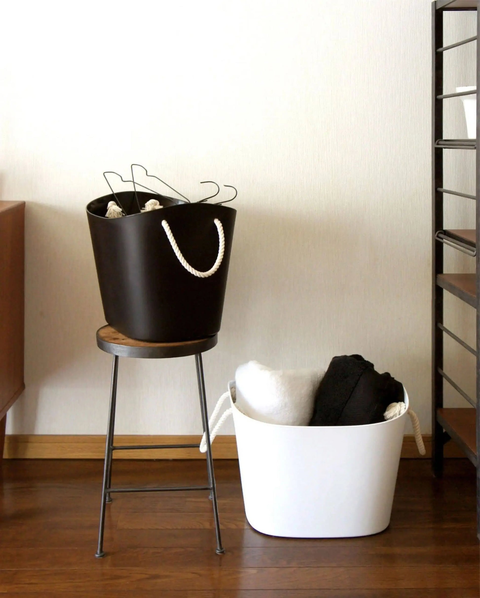 A black Hachiman Balcolore Storage Basket M with rope handles holds hangers on a wooden stool next to a white storage bin of clothes, all arranged on a wooden floor for a minimalist Japanese look against a plain wall.