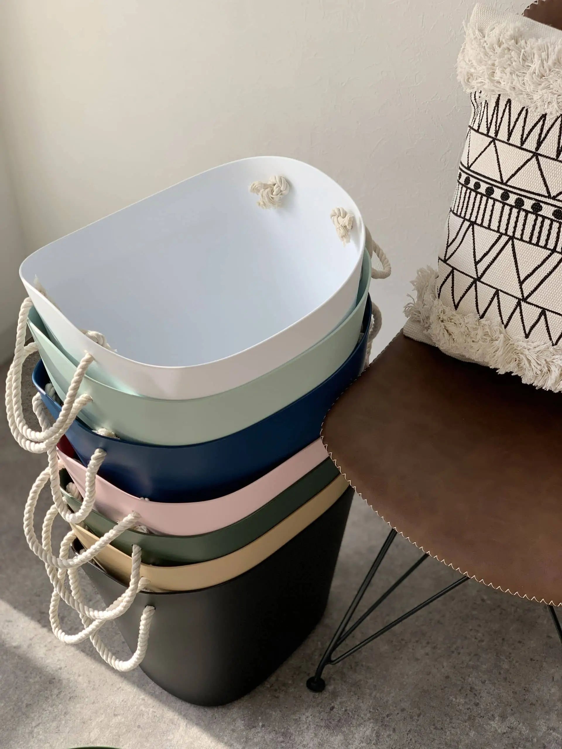 A stack of five Hachiman Balcolore Storage Basket M in pastel colors with rope handles sits beside a brown chair with a geometric cushion, bathed in natural light—ideal for minimalist Japanese-style multi-purpose storage.