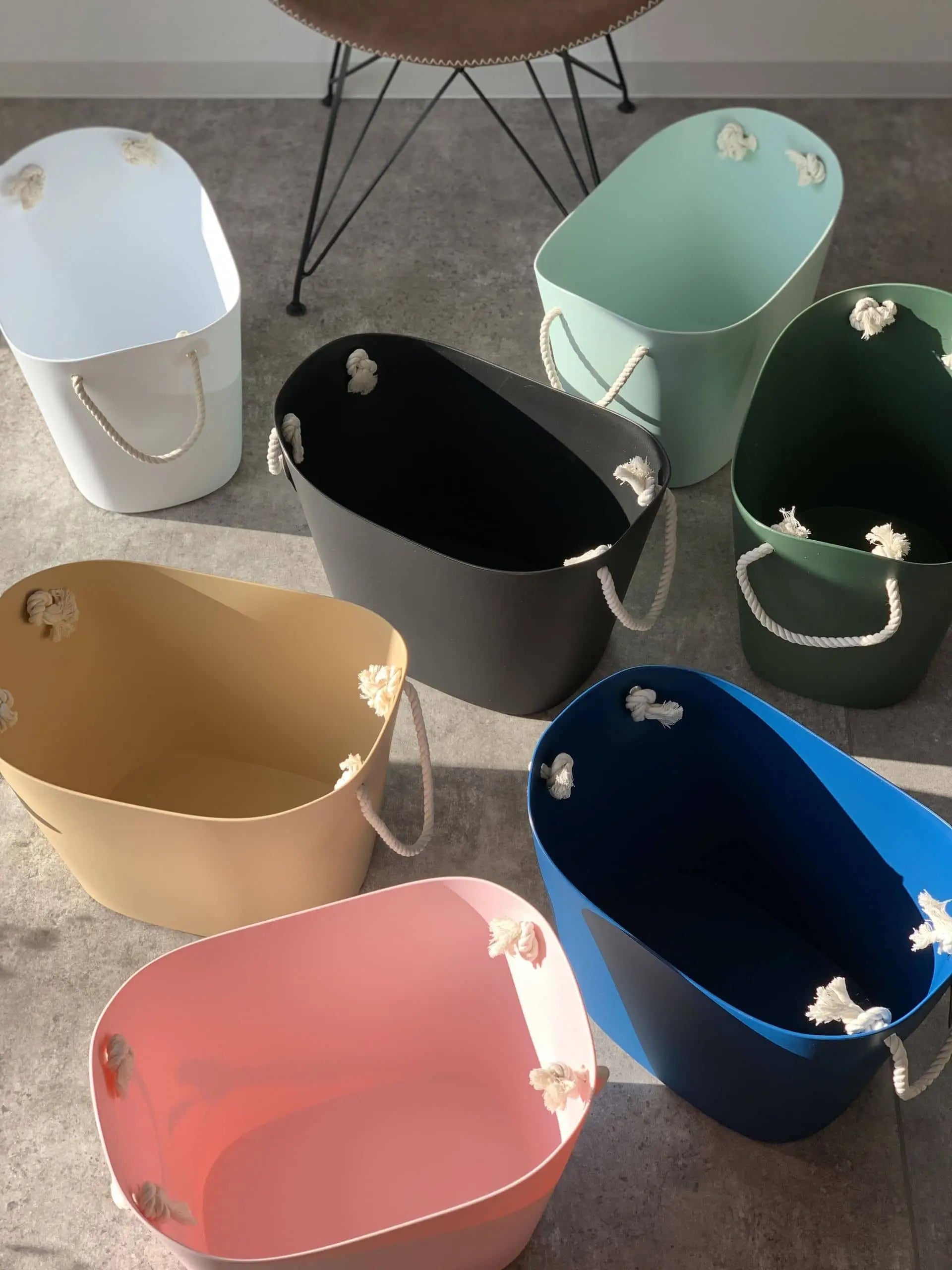 Seven Hachiman Balcolore Storage Basket M containers in white, black, green, yellow, pink, blue, and light green with rope handles sit on a gray floor, showcasing minimalist Japanese design and versatile storage.