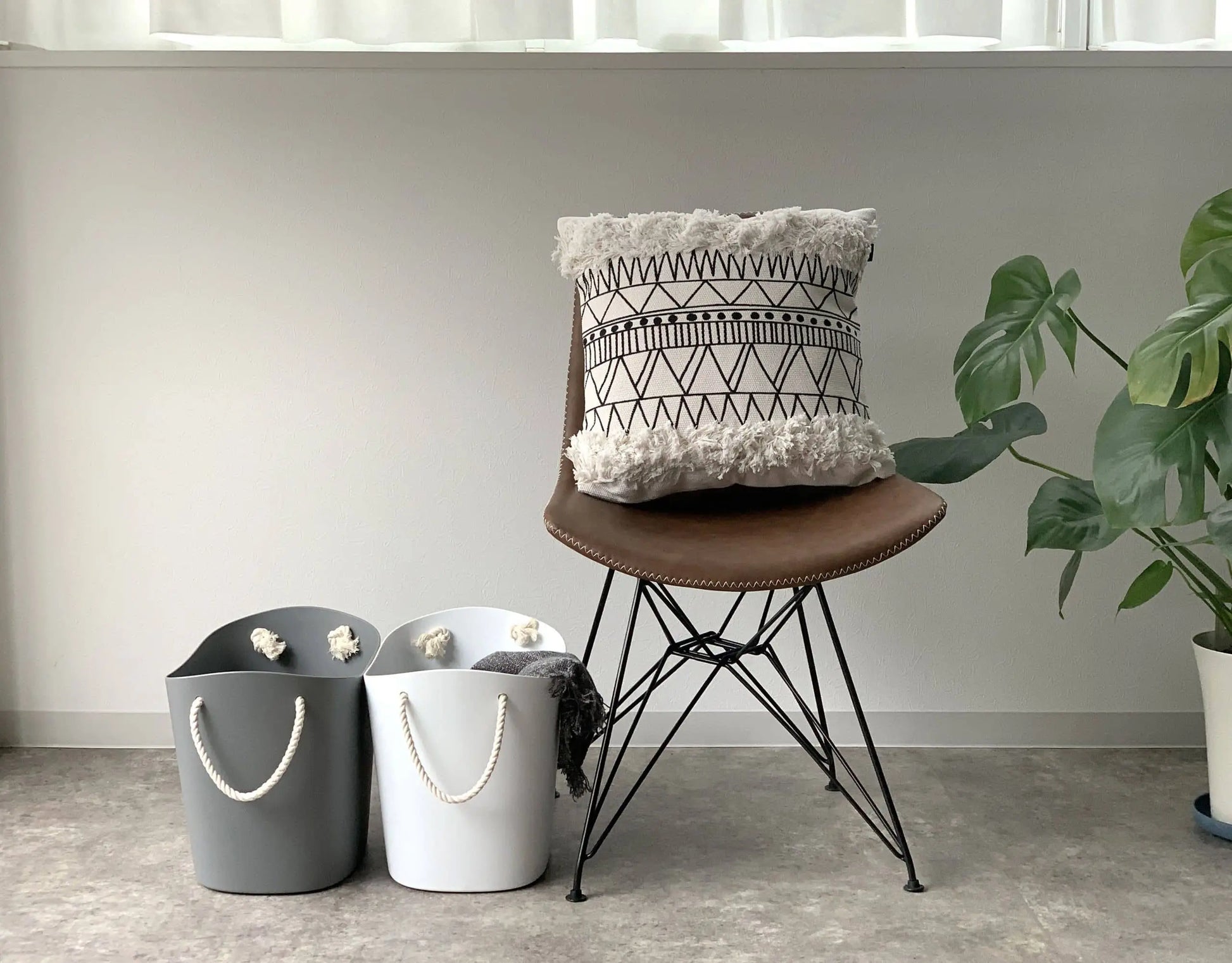 A modern brown chair with a patterned cushion sits beside a green potted plant. Two Hachiman Balcolore Storage Basket M, known for minimalist Japanese style and versatile storage, are placed on the floor against a white wall.