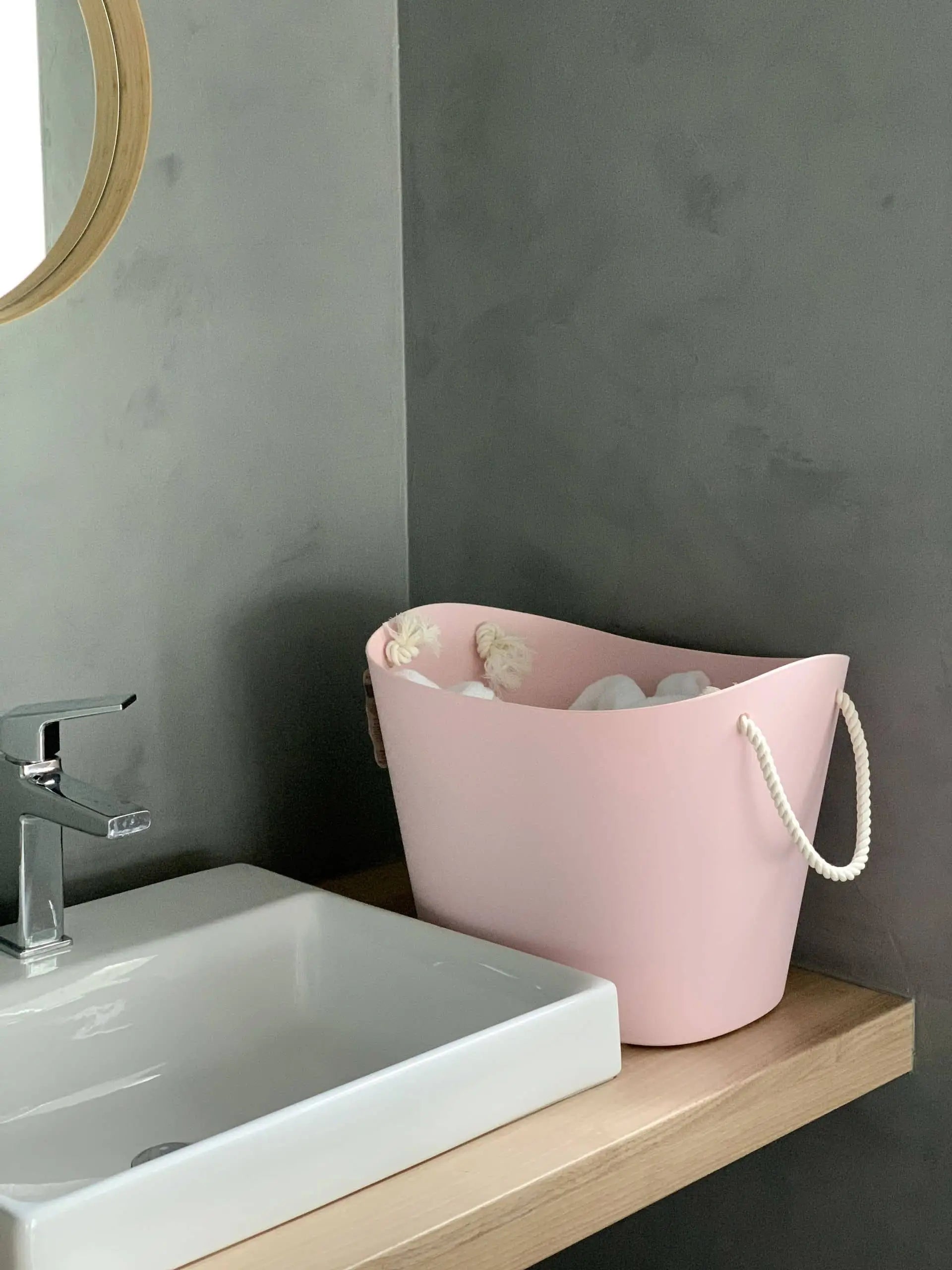 A pink Hachiman Balcolore Storage Basket M with rope handles sits on a wooden countertop by a white sink in a gray-walled, minimalist Japanese bathroom—ideal for stylish, multi-purpose storage.