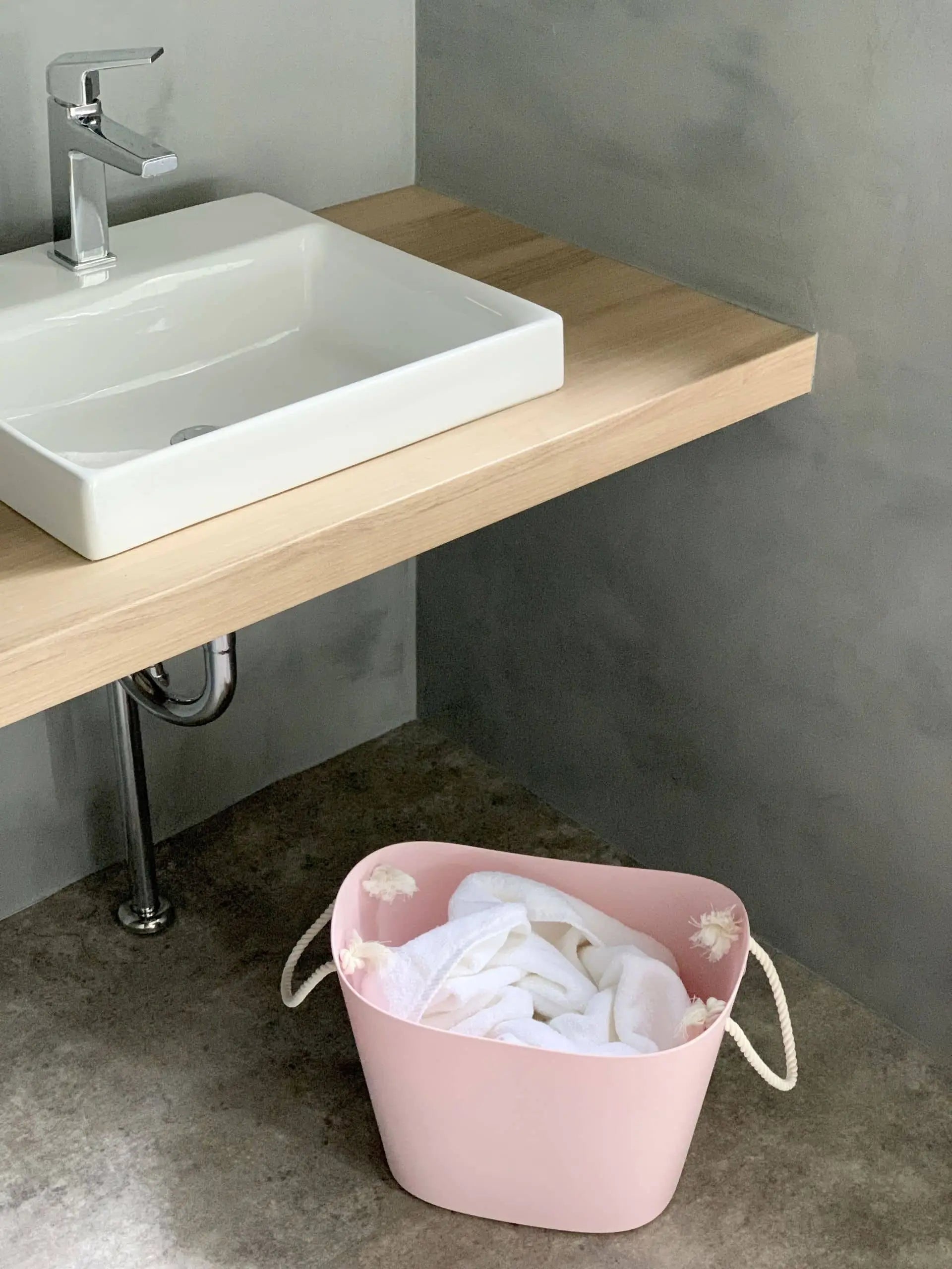 A Hachiman Balcolore Storage Basket M in pink with white handles, filled with white clothes, is placed on the floor under a wooden countertop with a modern white sink and silver faucet in a minimalist Japanese bathroom.