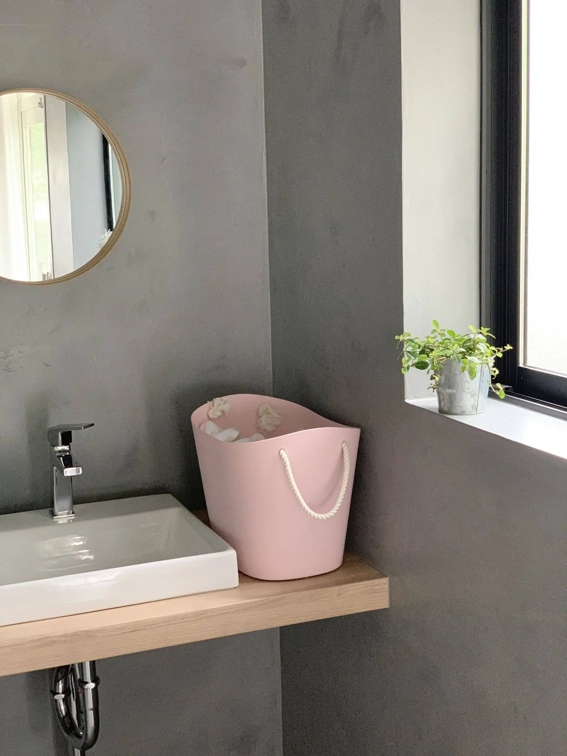 A modern bathroom with a square white sink, a round mirror on a gray wall, and a Hachiman Balcolore Storage Basket M by Hachiman with white rope handles on the wooden counter, plus a small potted plant on the windowsill.