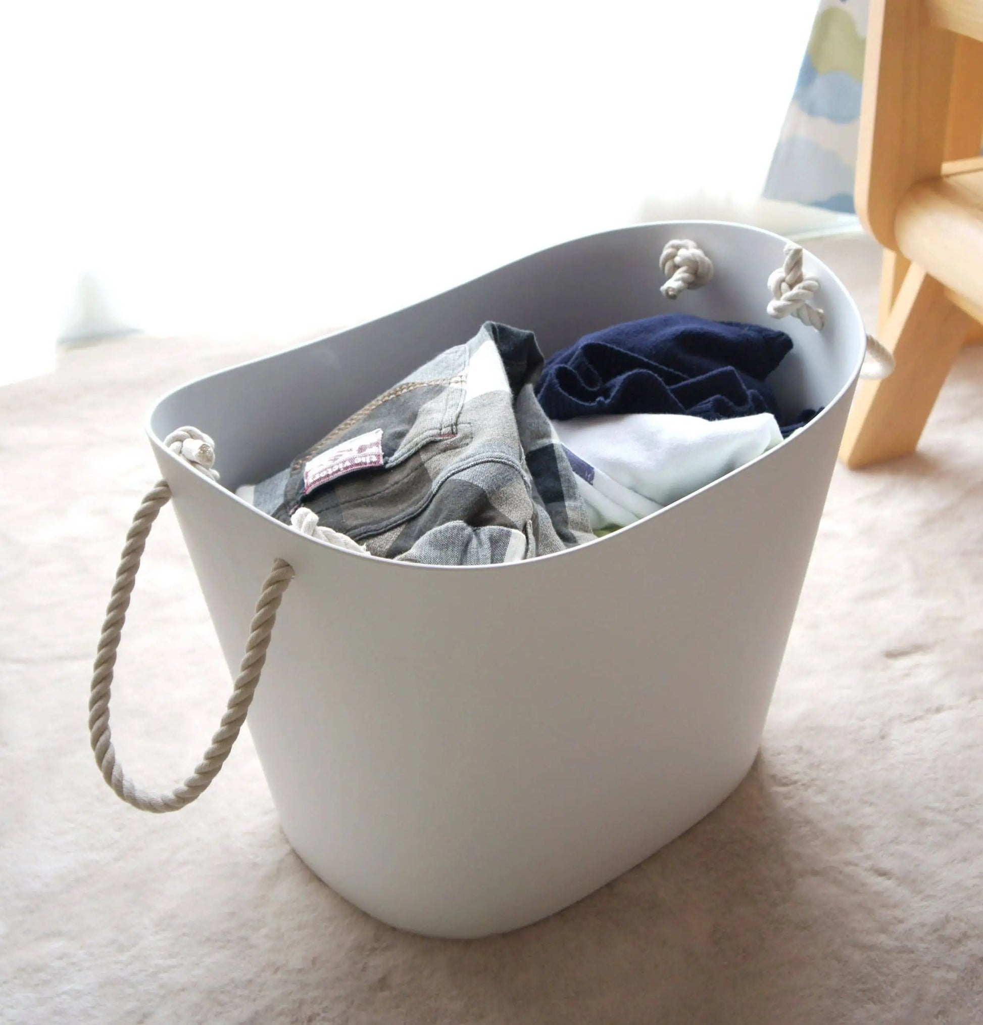 A Hachiman Balcolore Storage Basket M with rope handles, filled with folded clothes, rests on a light carpet near a window and wooden furniture—perfect for minimalist Japanese or versatile storage by Hachiman.