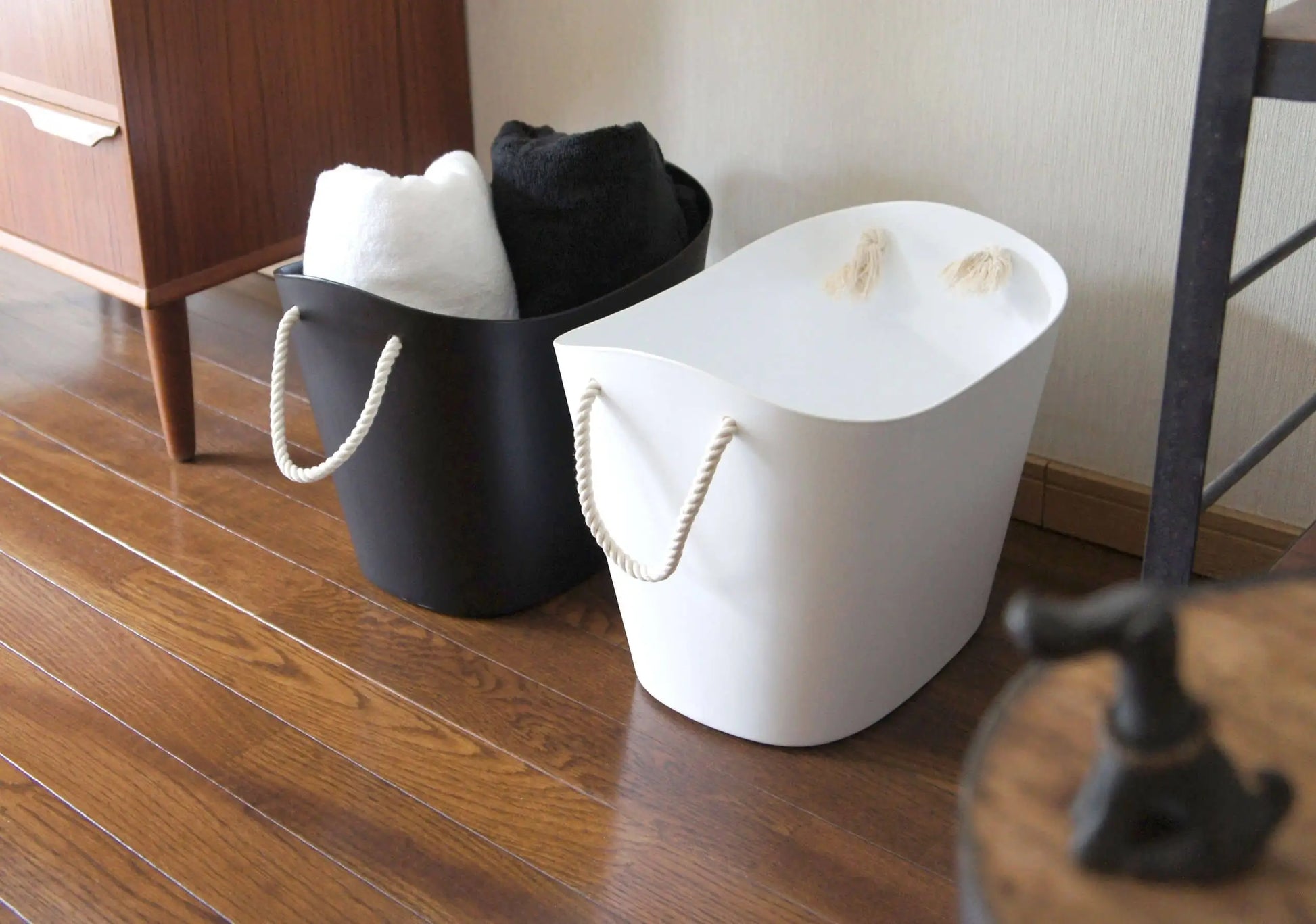 Two Hachiman Balcolore Storage Basket M in black and white with rope handles sit on a wooden floor. Ideal for minimalist Japanese spaces, the black holds rolled towels while the white offers versatile and multi-purpose storage.