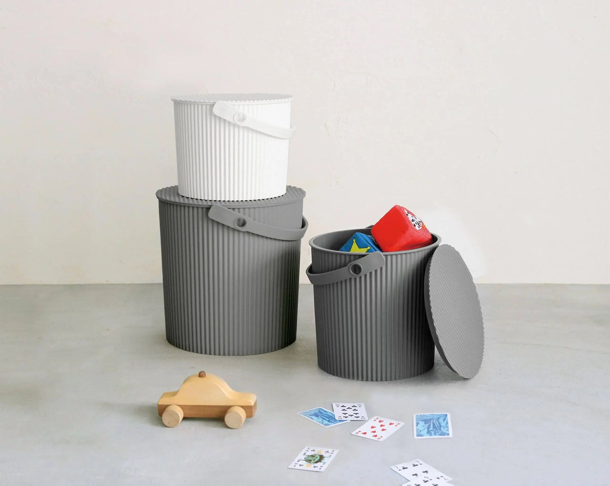 Three Hachiman Omnioutil Storage Bucket L bins—two gray and one white—are stacked on a light floor. The open gray bin holds toys; a wooden toy car and cards are scattered nearby. Durable Hachiman design, perfect for indoor and outdoor use.