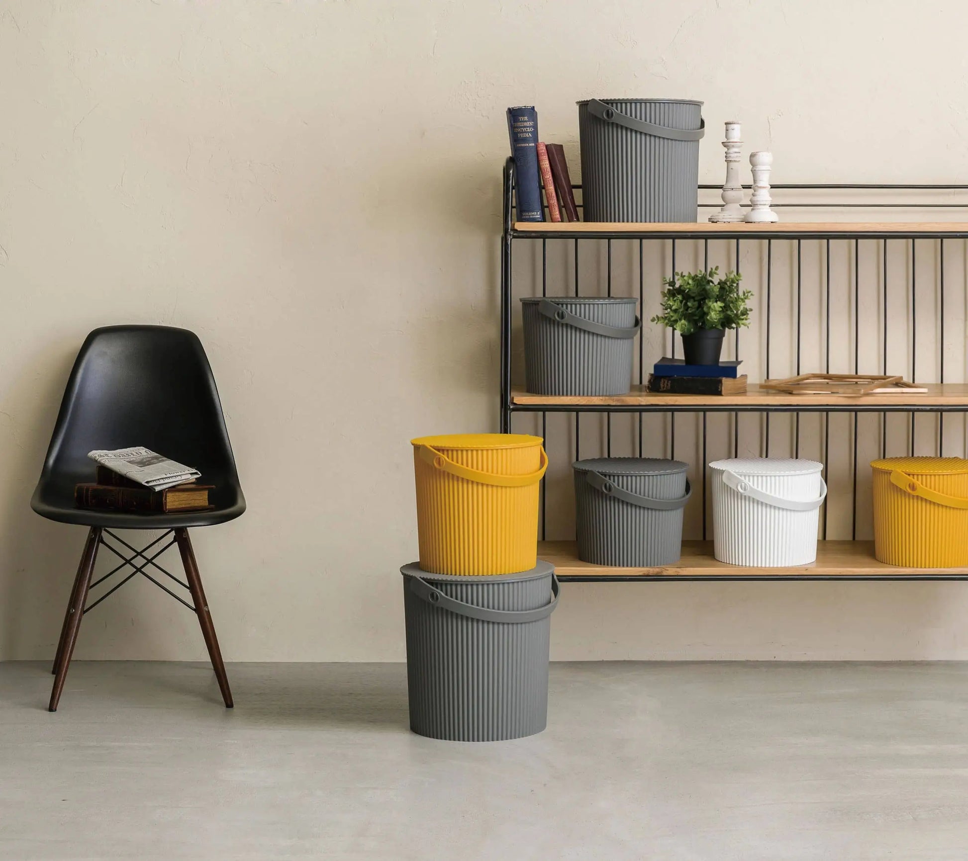 A modern interior features a black chair with books and a shelf displaying colorful Hachiman Omnioutil Storage Bucket S by Hachiman, books, potted plants, and white candleholders against a light wall.