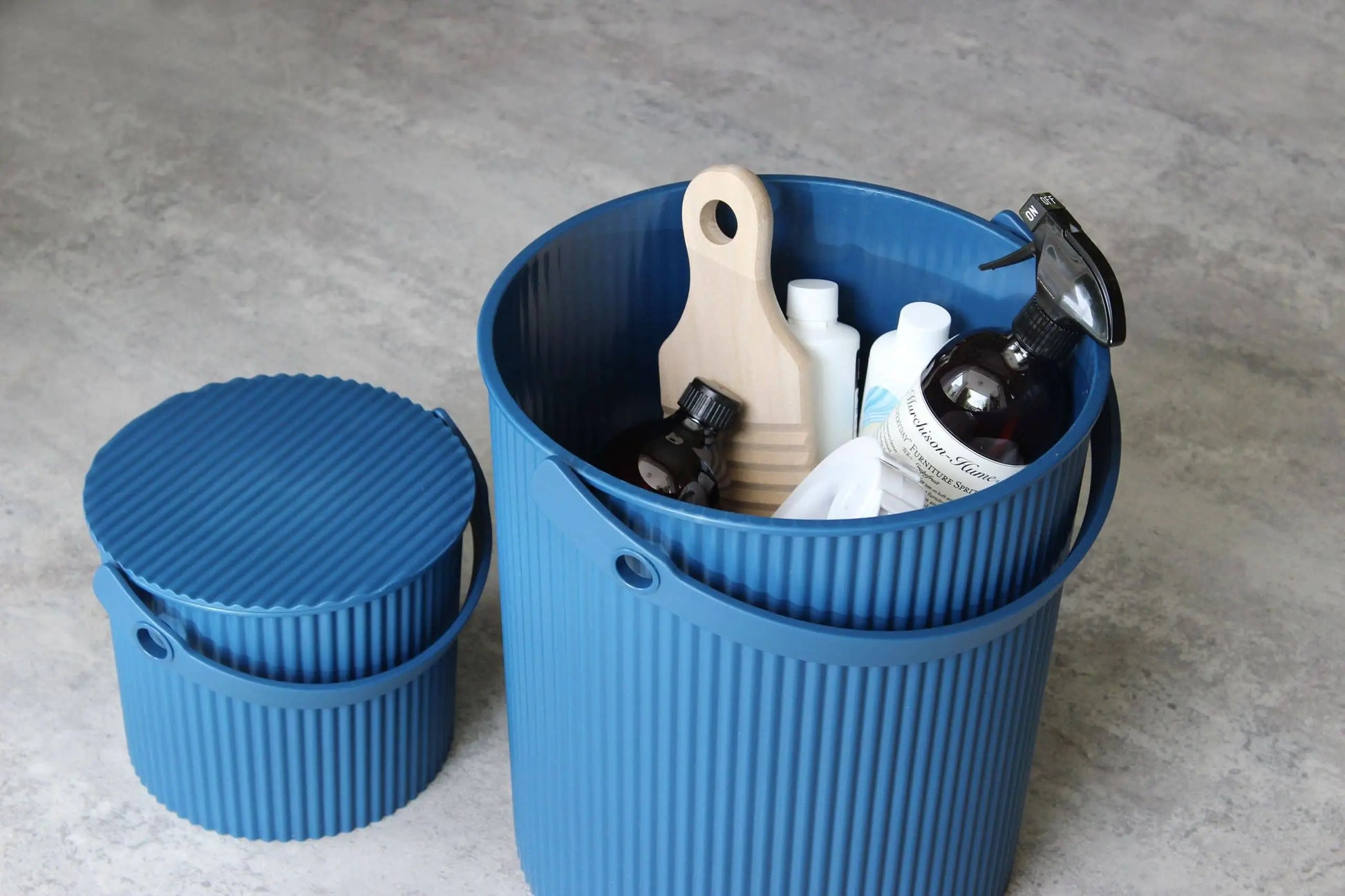 Two ribbed blue Hachiman Omnioutil Storage Bucket S bins sit on a gray floor—one holds cleaning supplies with the lid off, the other is closed. Durable and versatile, Hachiman buckets are ideal for indoor or outdoor storage needs.