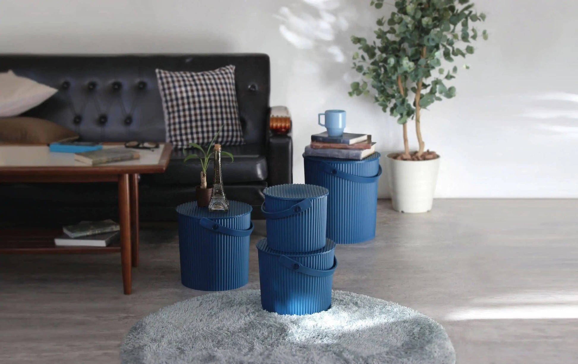 A modern living room with a black sofa, checkered cushion, wooden coffee table, three stacked Hachiman Omnioutil Storage Buckets S by Hachiman for indoor/outdoor use, a potted plant, and books on a light gray rug and floor.