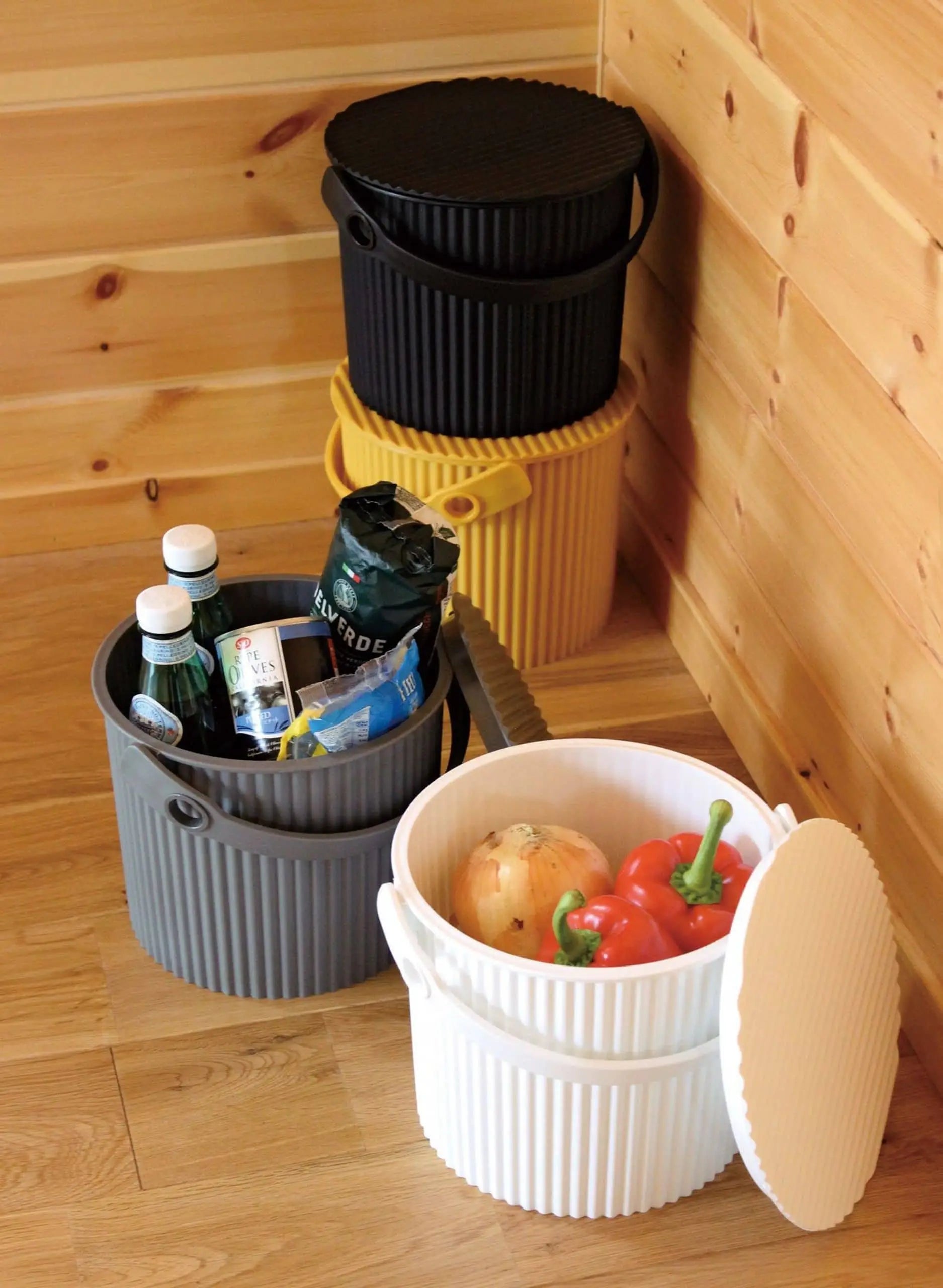 Four ribbed Hachiman Omnioutil Storage Bucket S containers by Hachiman, in black, yellow, gray, and white, sit on a wooden floor. Two are open—one holding drinks and snacks, the other an onion and red bell peppers. Ideal for indoor or outdoor use.