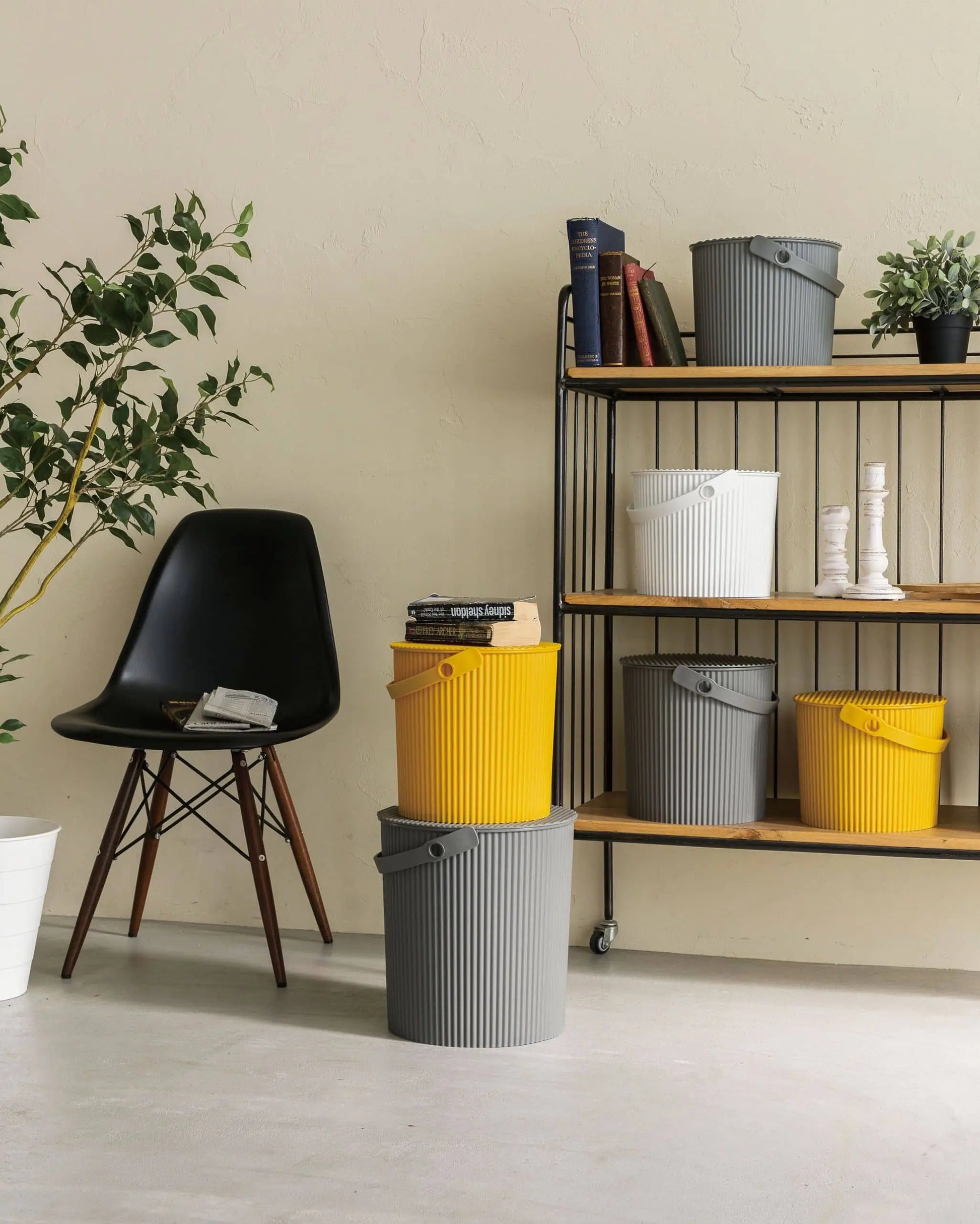 A modern room features a black chair, potted plant, and books on a metal shelf with Hachiman Omnioutil Storage Bucket S in white, yellow, and gray by Hachiman. Two more buckets are stacked on the floor beside the chair for indoor or outdoor use.