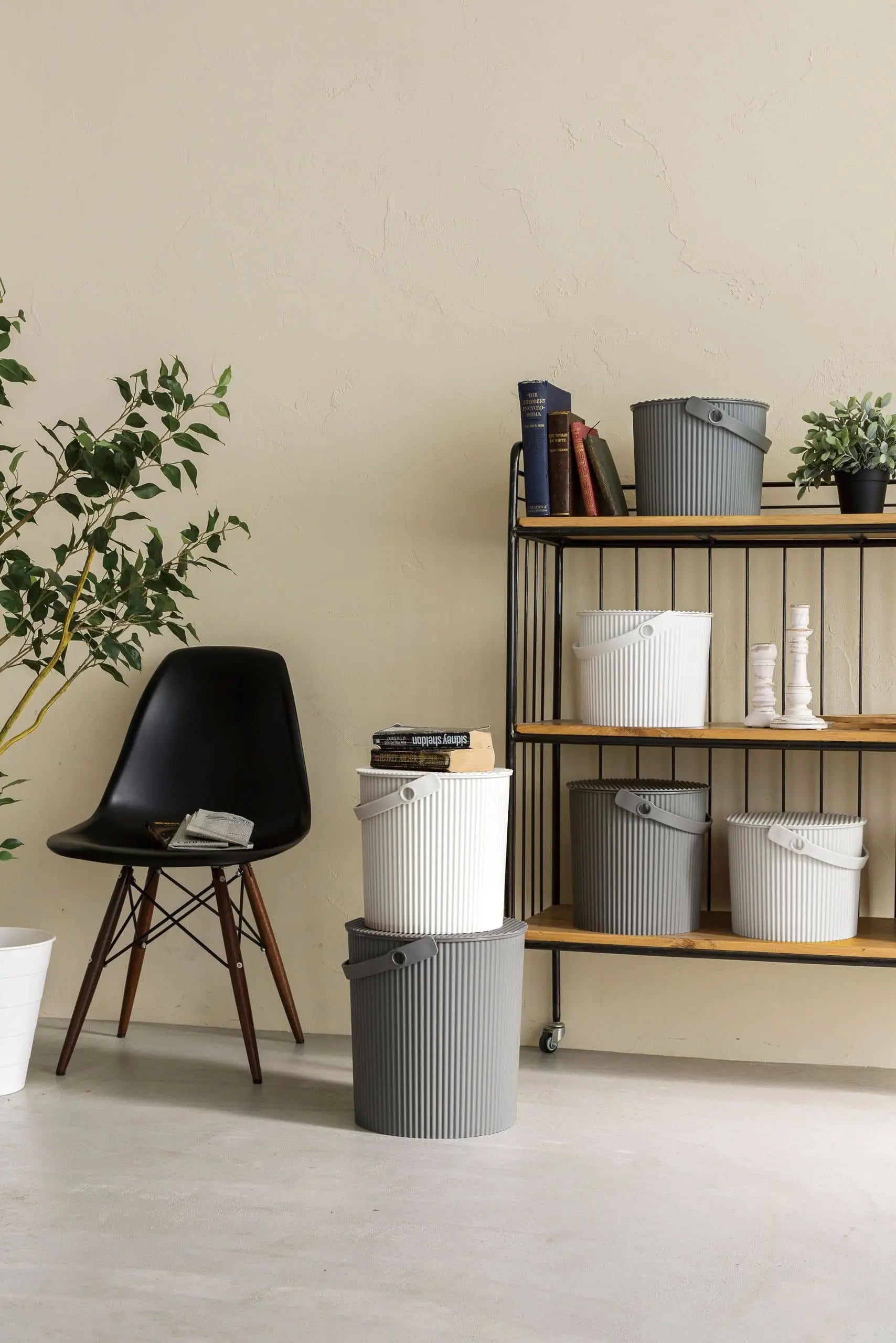 A modern interior with a black chair beside stacked gray and white Hachiman Omnioutil Storage Bucket S. A metal and wood shelf displays more Hachiman buckets, books, and potted plants. A leafy plant sits to the left against a beige wall.