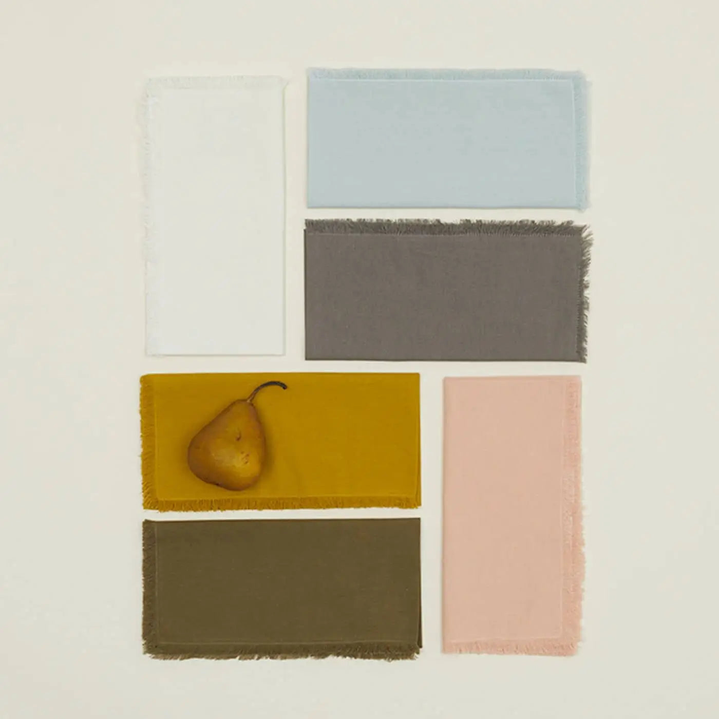 Six rectangular fabric swatches, including the mustard yellow Hawkins New York Essential Dinner Napkin (Set of 4), are arranged in a grid on a light background, with a pear resting on the dinner napkin in the lower left section.