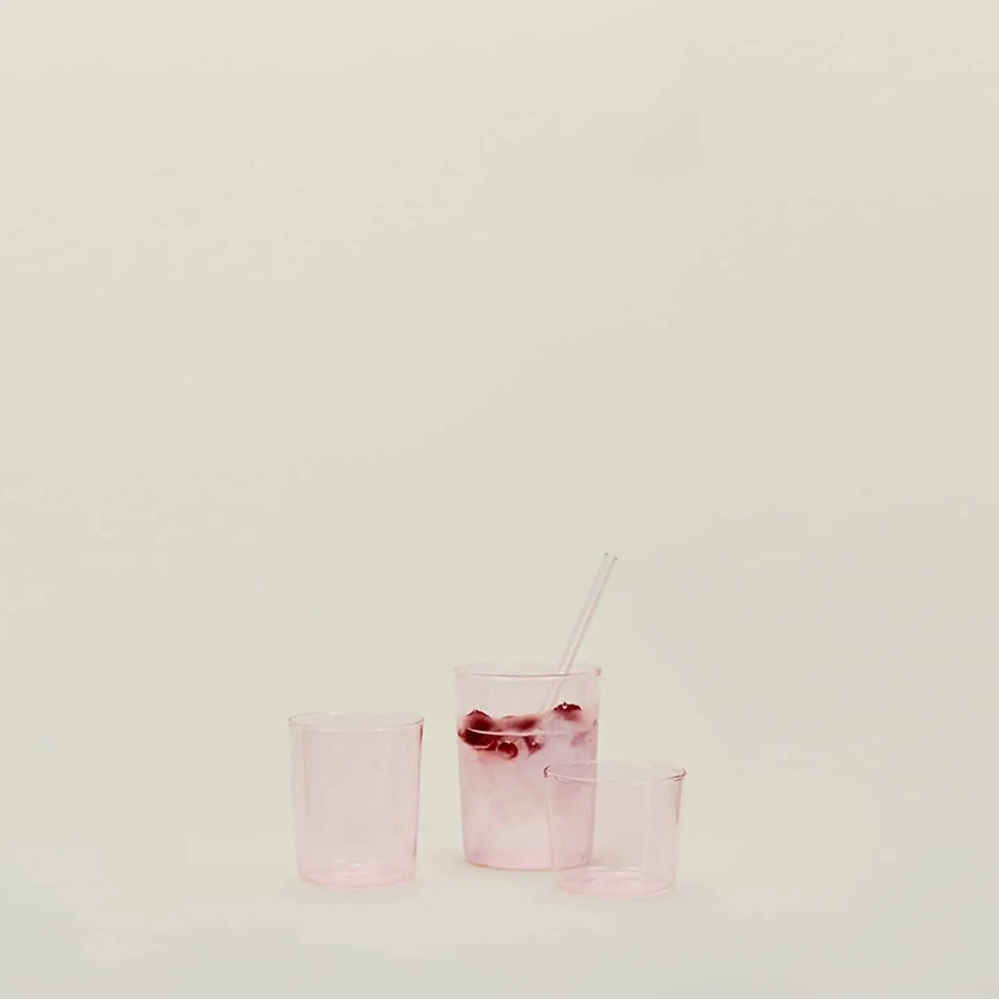 Three translucent pink Hawkins New York Essential Glassware, Set Of 4, Medium glasses sit on a plain light surface. The tallest holds ice, a clear straw, and pink liquid; the other two remain empty against a neutral background.