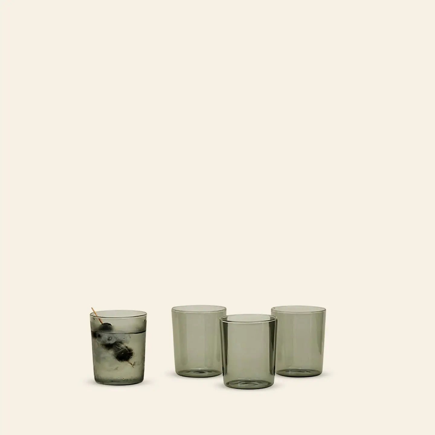 A set of four Hawkins New York Essential Glassware, Medium, sits on a plain surface; one glass is filled with ice and a drink with a cocktail pick, while the other three remain empty against a beige background.