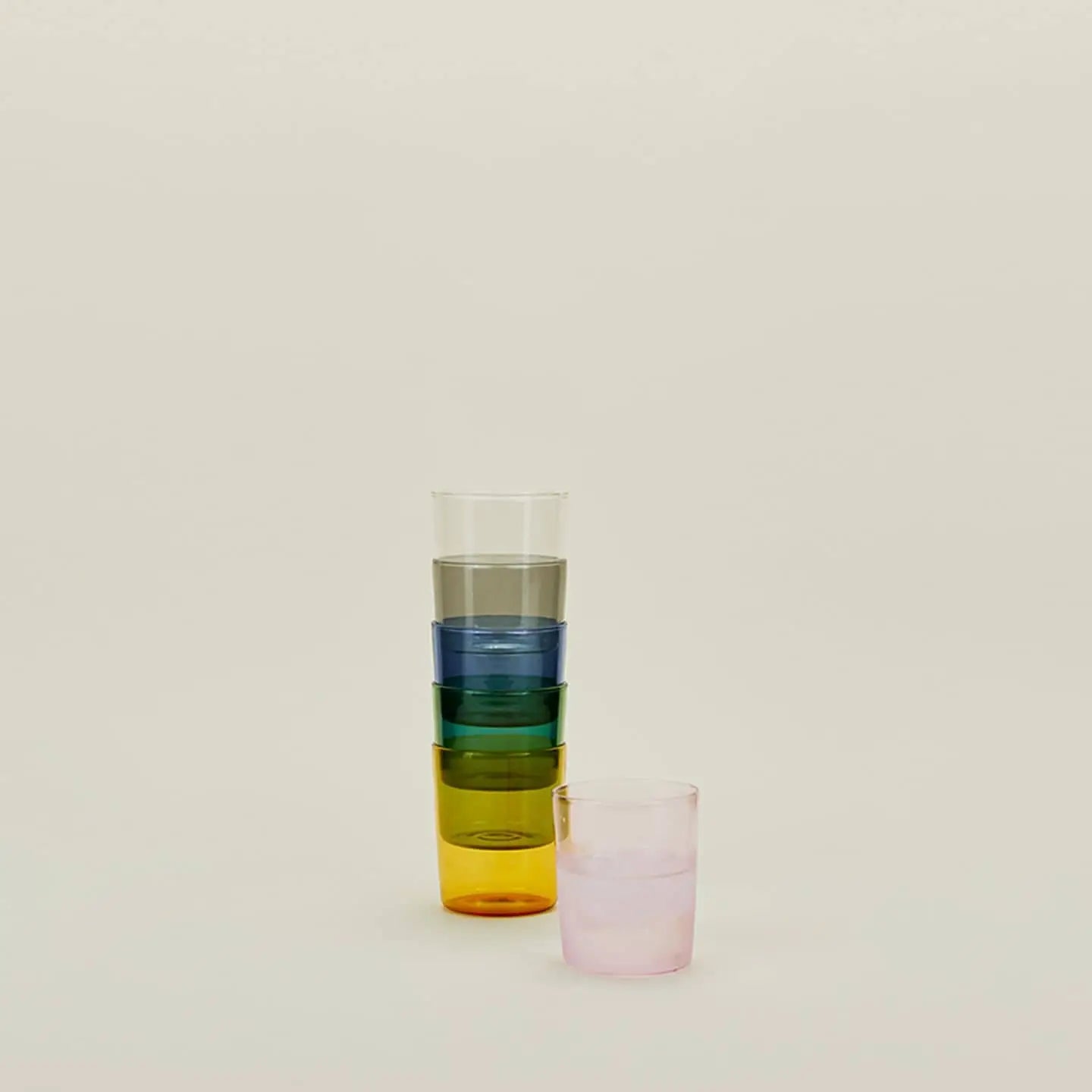 Four Hawkins New York Essential Glassware cups—clear, blue, green, and yellow—are stacked vertically, with a pink cup in front. The set of 4 medium cups is displayed against a plain beige background.