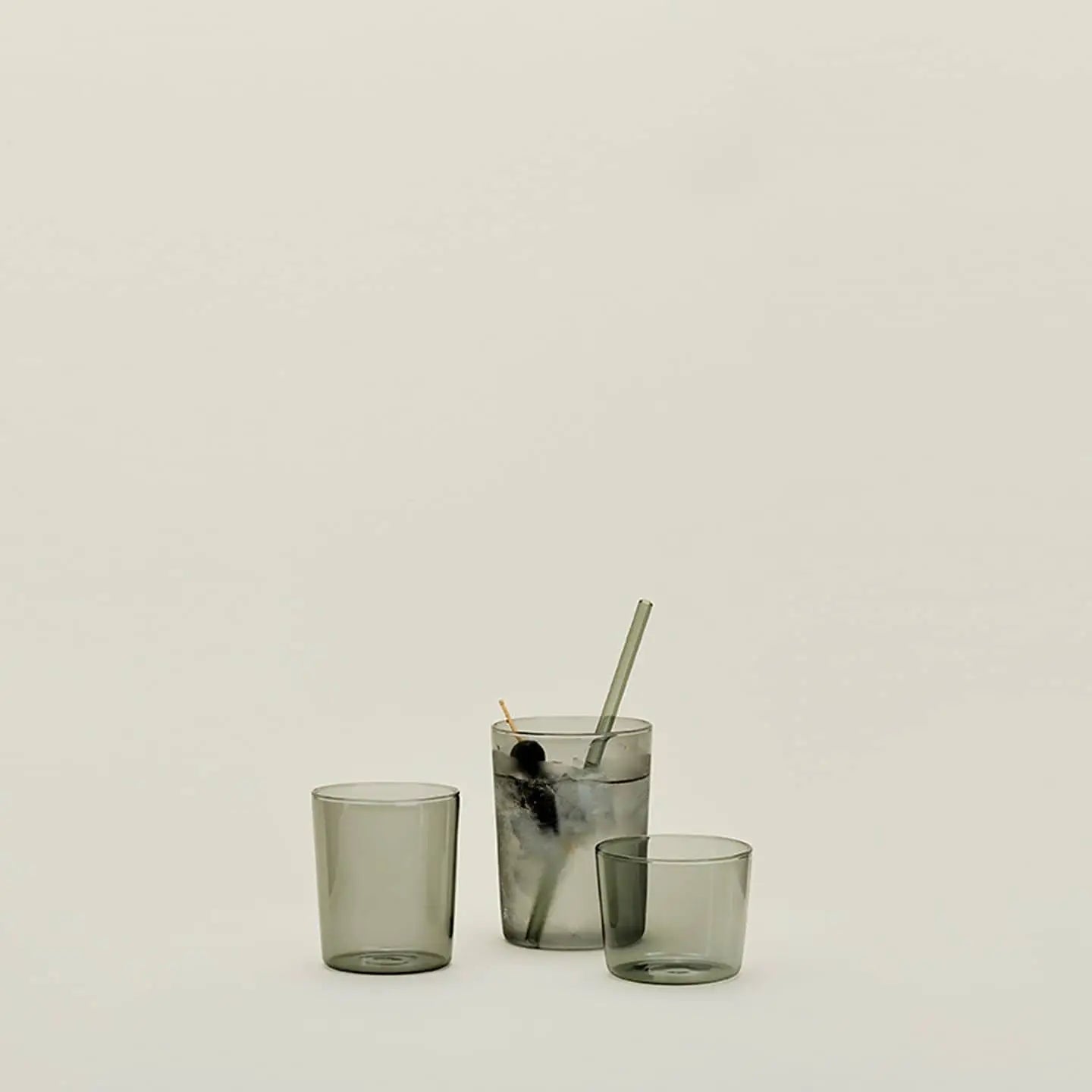 Three Hawkins New York Essential Glassware medium drinking glasses sit on a plain light background; one, highlighting quality glass construction, holds ice, a dark cherry, a metal straw, and cocktail pick while the other two remain empty.