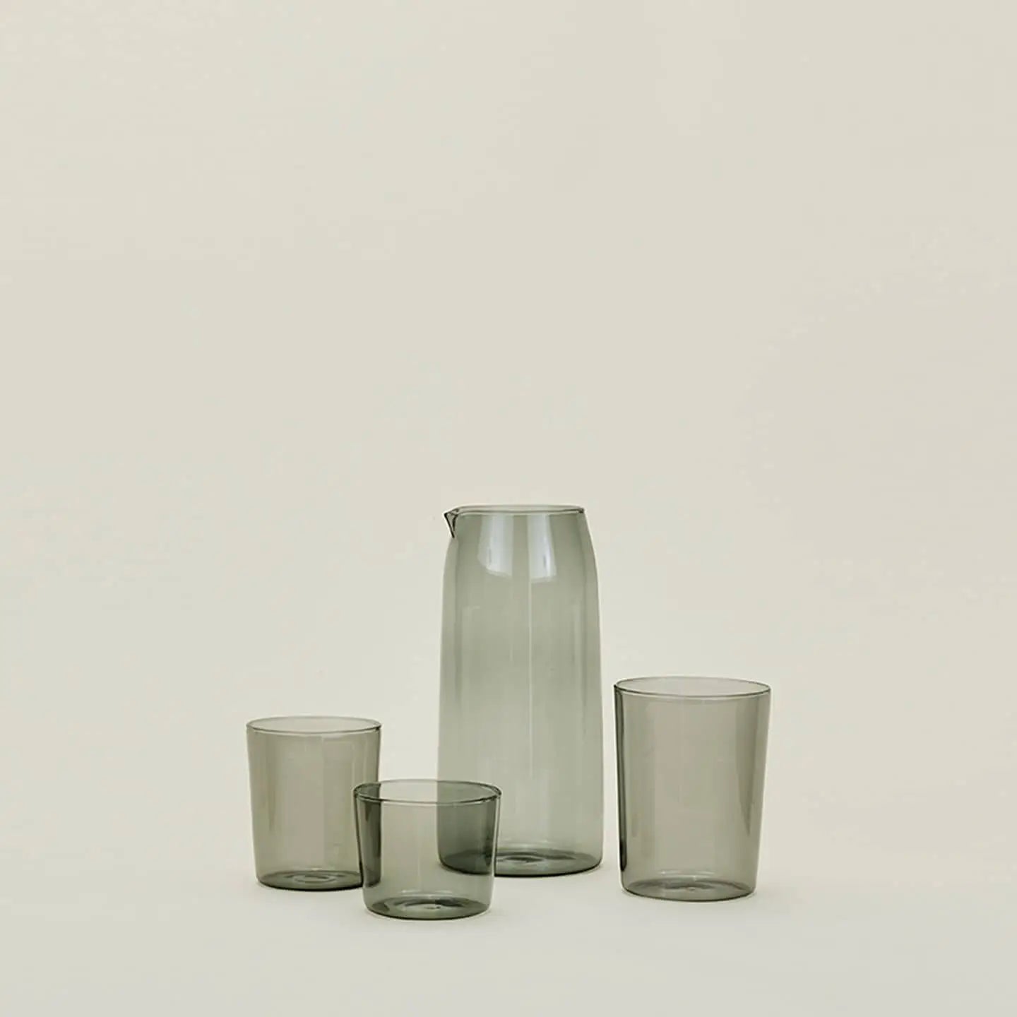 The Hawkins New York Essential Glassware, Set Of 4, Medium features one tall carafe and three drinking glasses of varying heights in translucent gray glass, highlighting premium construction against a light beige background.