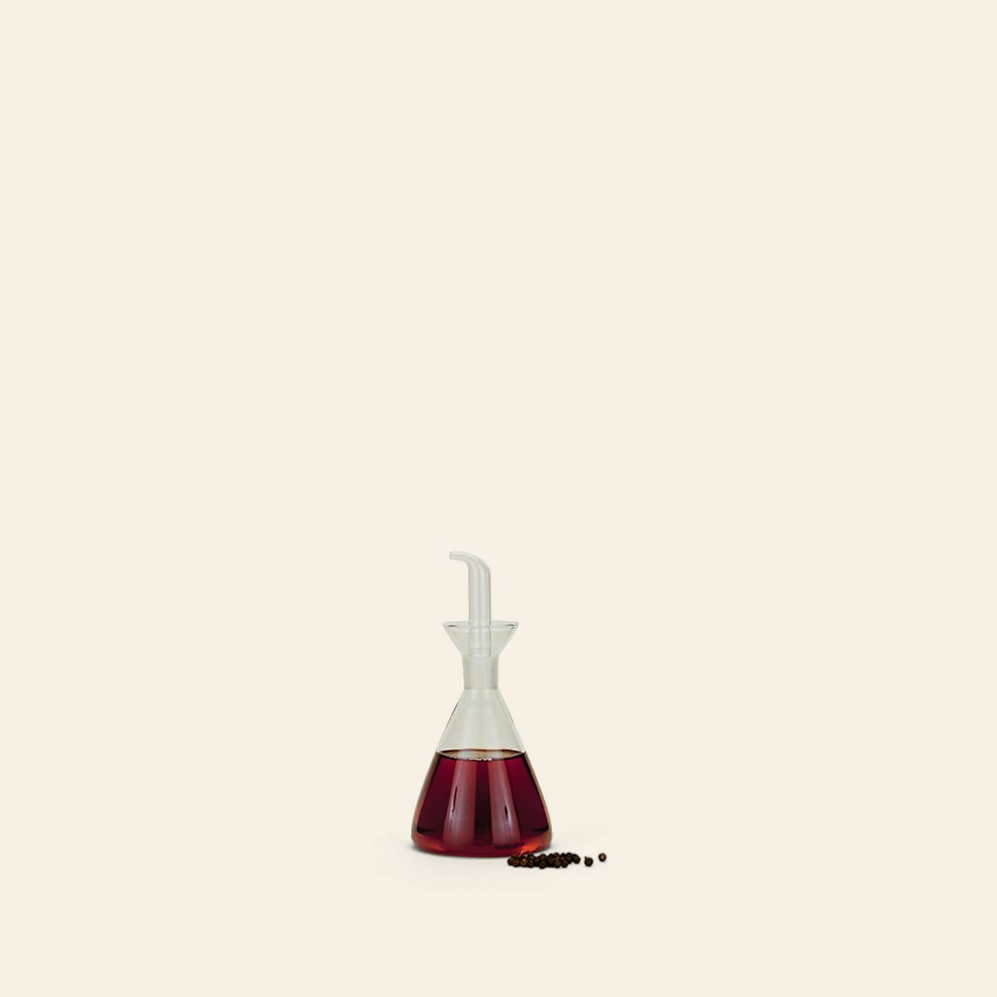 The Hawkins New York Essential Kitchen Bottle, Medium, with modern styling and a precision drizzle spout, holds red liquid—possibly vinegar—and sits on a light surface beside peppercorns against a pale background.