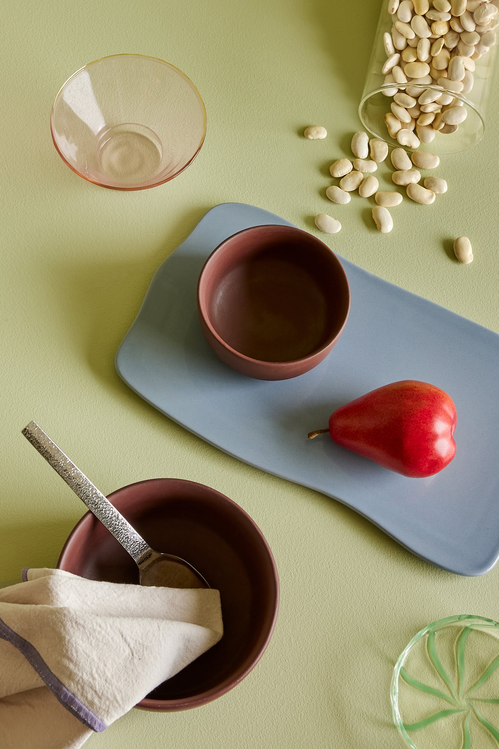 A red pear on a blue tray sits beside the Hübsch Clay Bowls, Set of 2 by Hübsch—versatile bowls paired with a cloth and spoon—a glass bowl, and scattered white beans on a light green surface.
