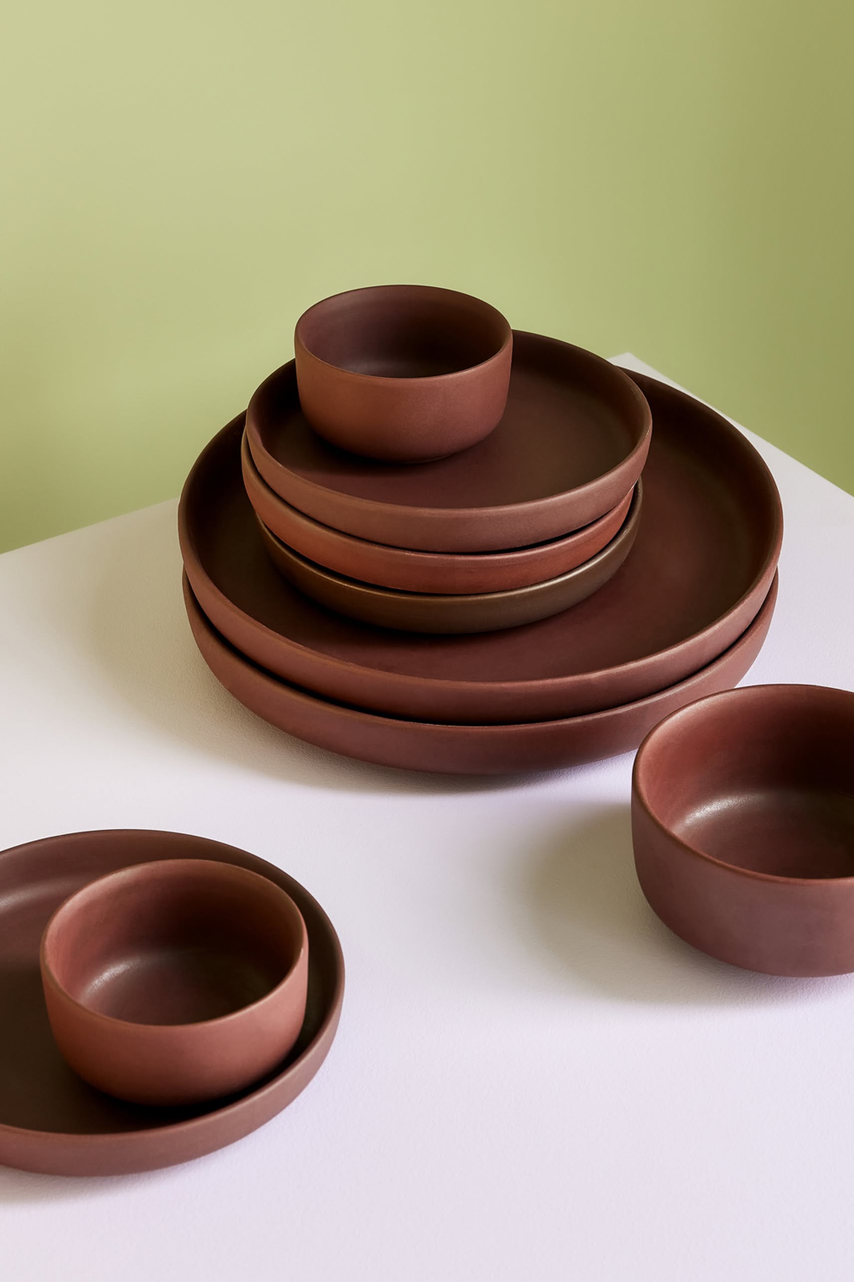 The Hübsch Clay Bowls, Set of 2 by Hübsch features matte brown bowls and plates with an elegant design, neatly stacked on a white surface against a green backdrop, with some versatile pieces displayed separately in front.