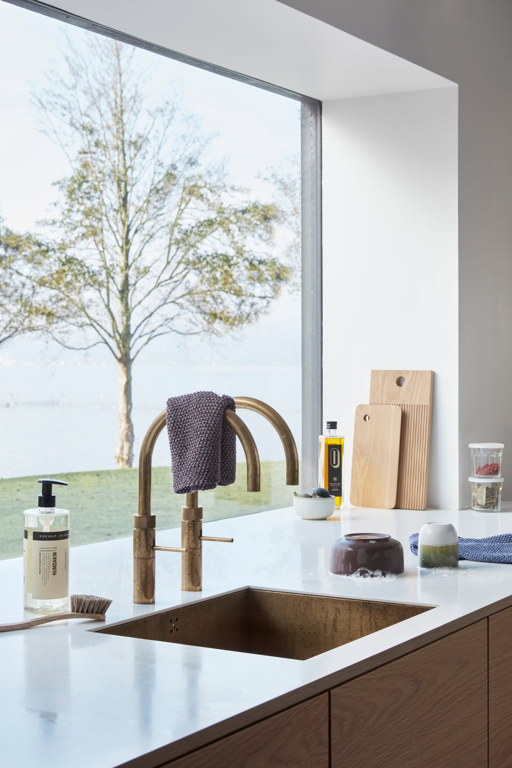A modern kitchen sink with a brass faucet sits beneath a window. On the countertop, Hübsch Clay Bowls, Set of 2 by Hübsch join soap, towels, and cutting boards, accenting the neat and elegant design of this bright space.