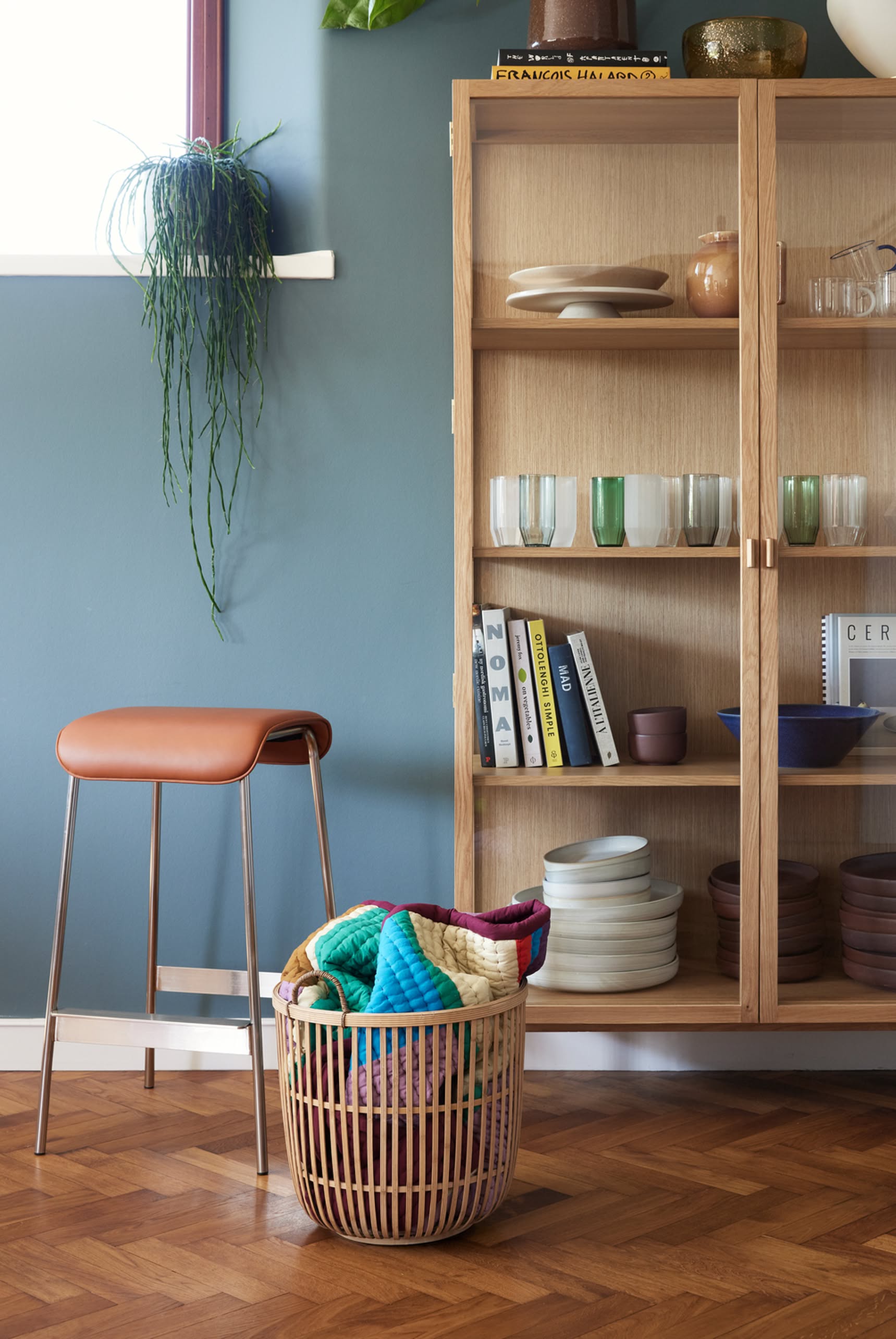 A wicker basket with blankets sits on a wood floor next to a tan-cushioned stool and a cabinet showcasing Hübsch Clay Bowls, Set of 2 by Hübsch, along with elegant dishes, glasses, and books against a blue wall.