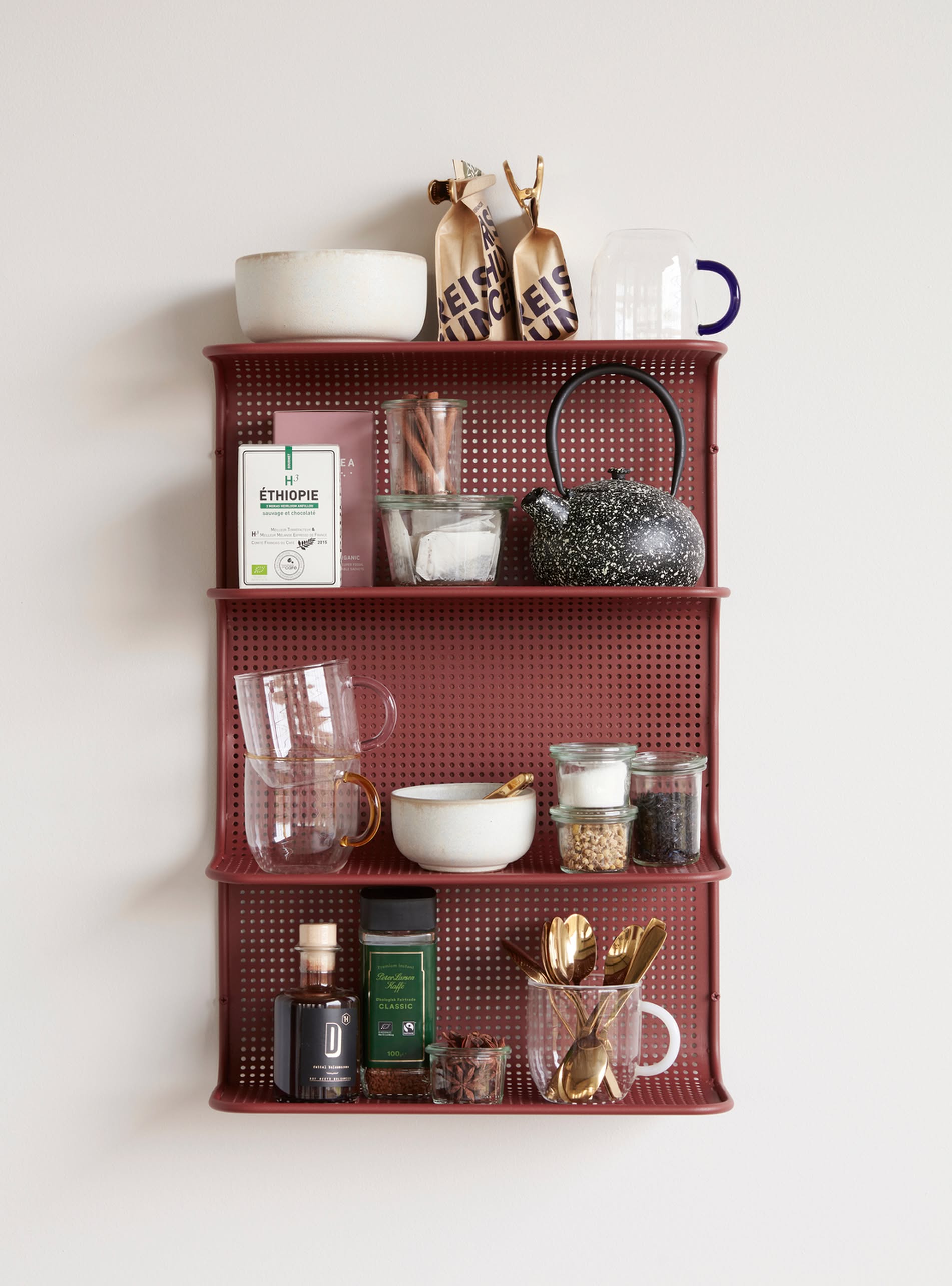 A red wall shelf displays coffee beans, a teapot, glasses, the Hübsch Clay Bowls Set of 2 by Hübsch, jars with spices, coffee and tea, mugs, and gold utensils—all neatly arranged against a white wall.