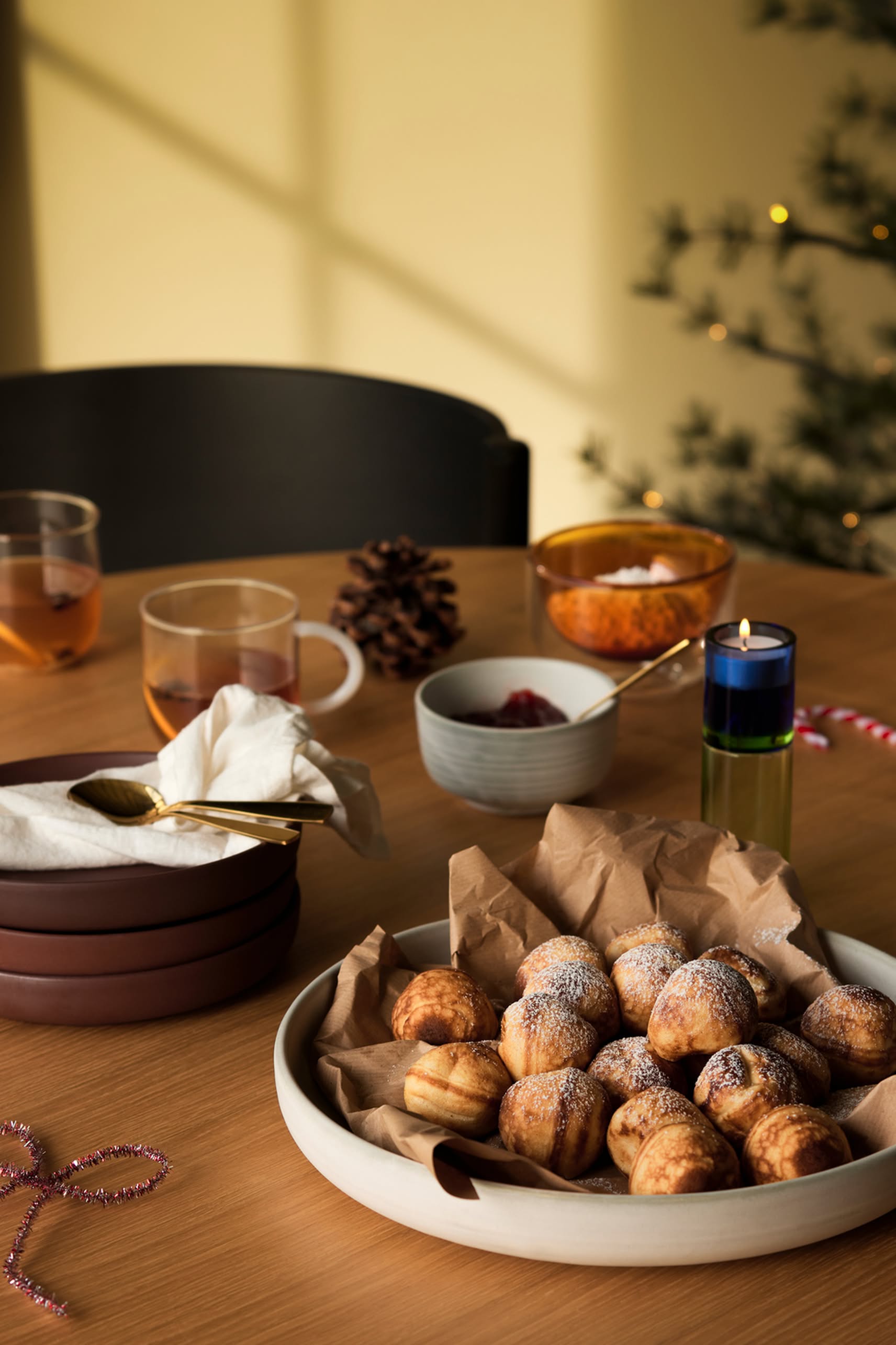A wooden table set with a plate of powdered pastries, the Hübsch Clay Bowls Set of 2 stacked with a napkin and spoon, cups of tea, small bowls of jam and orange spread, a pinecone, and a lit Christmas tree in the background.