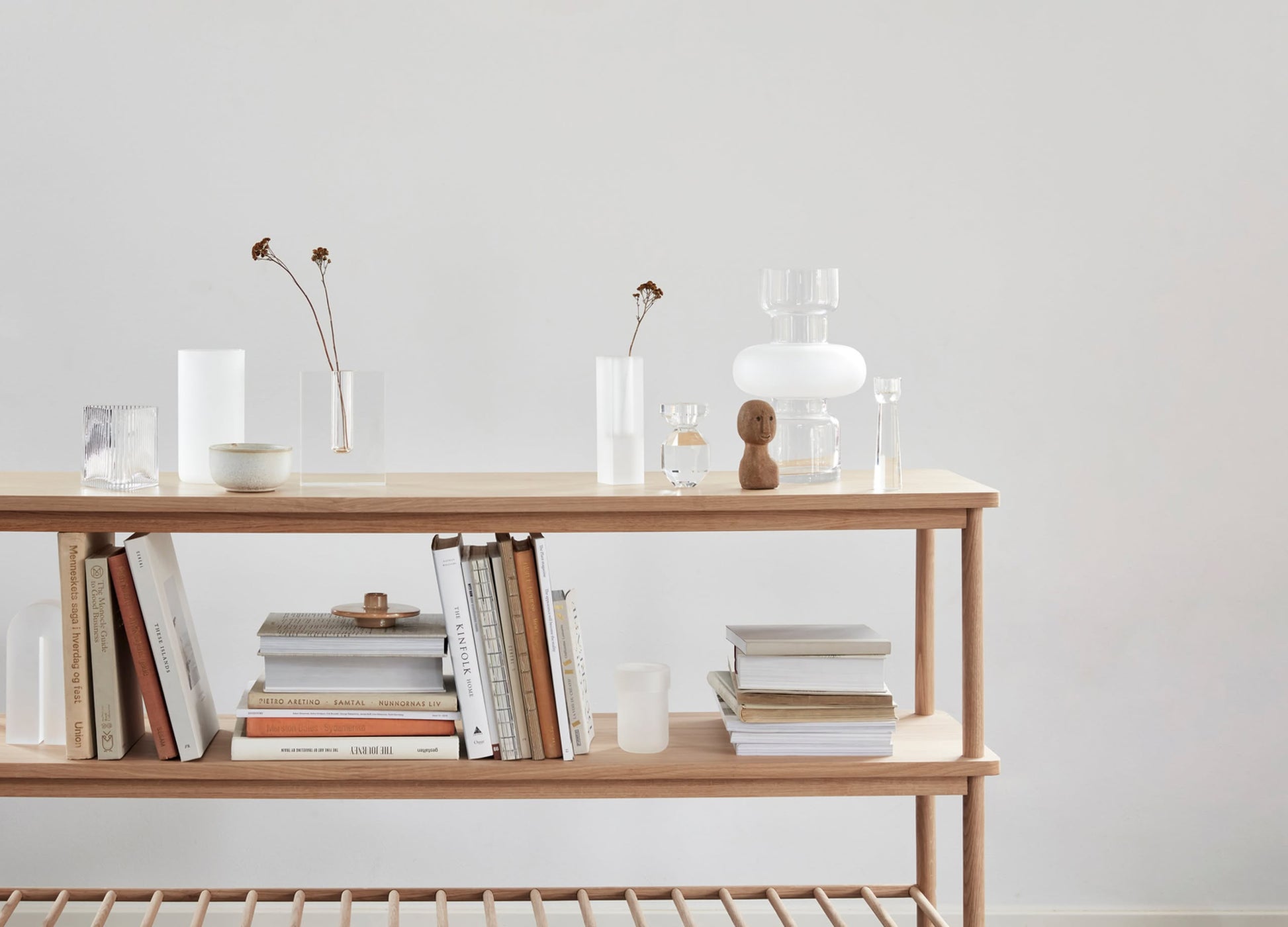 A minimalist wooden shelf against a plain light wall, styled with stacked books, elegant glassware, vases of dried flowers, a small wooden figurine, and a Hübsch Clay Bowls Set of 2 for refined charm.