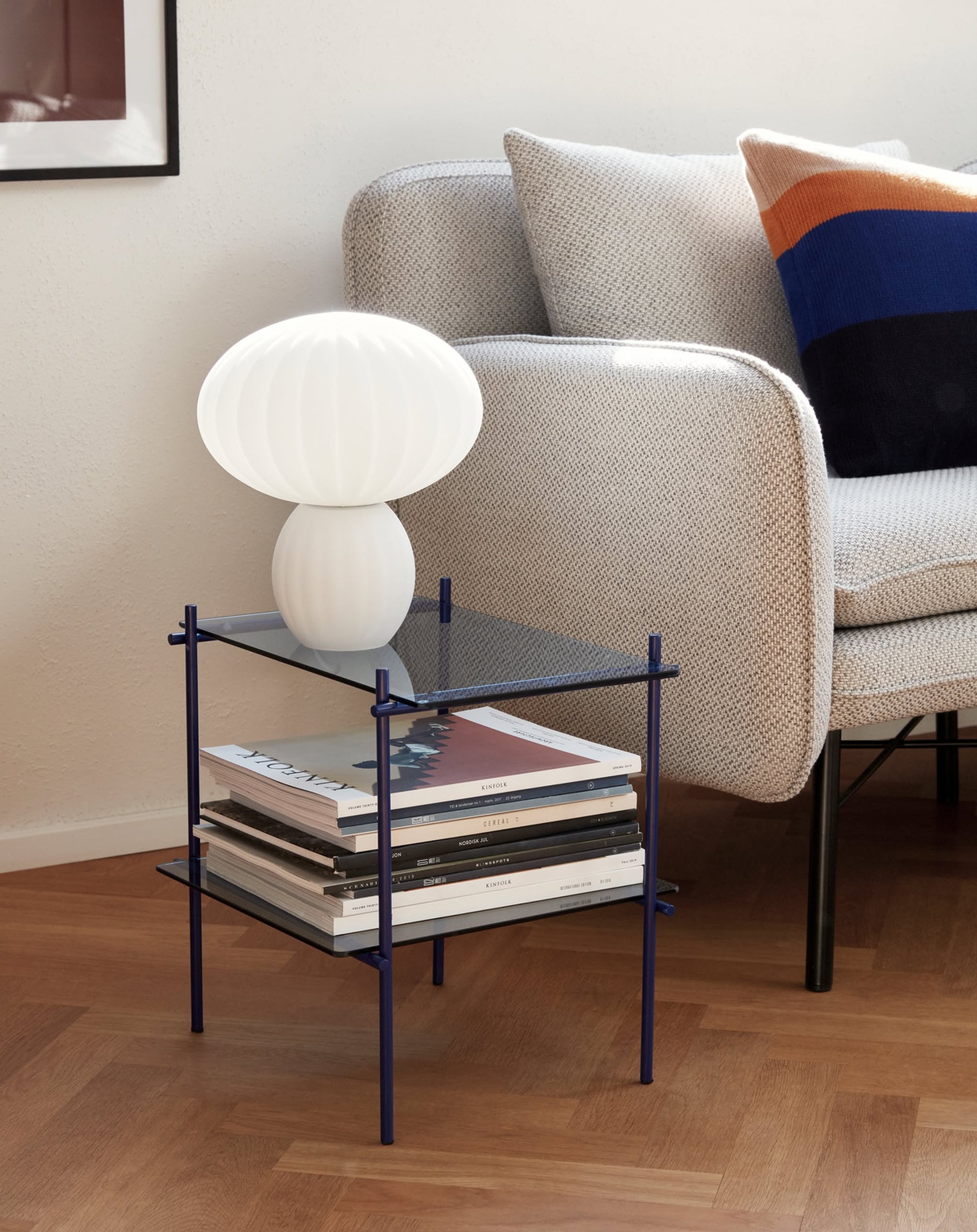 A modern living room corner features a light gray armchair, colorful pillow, and a glass side table displaying the Hübsch Kumu Table Lamp by Hübsch with dimmable function, alongside stacked magazines on a wooden floor.