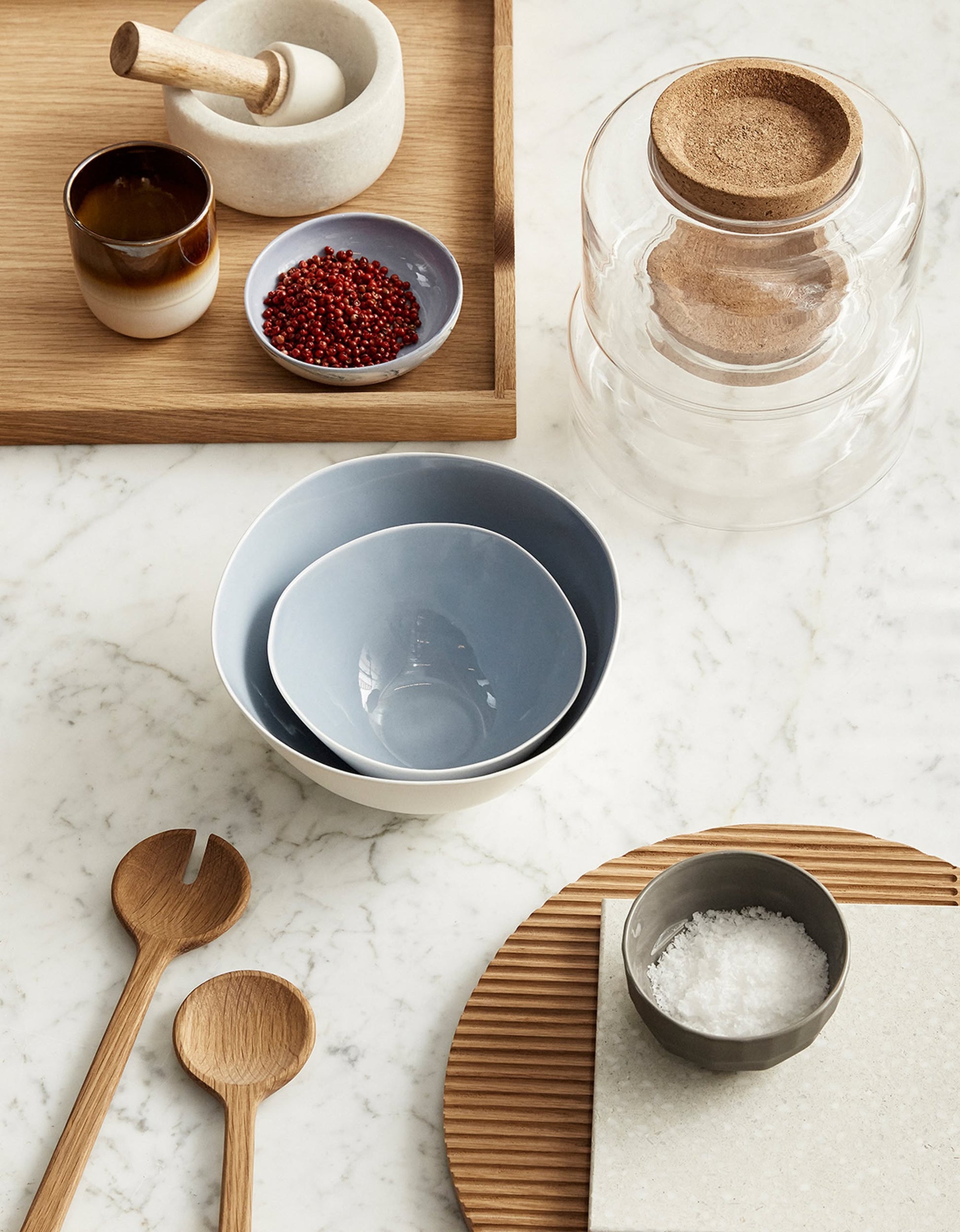 A marble countertop displays versatile kitchenware: the Hübsch Select Bowls, Set of 5 by Hübsch in nested blue porcelain. Also shown are a gray salt bowl, salad set, mortar and pestle, brown cup, peppercorns on a plate, and cork-lidded glass jar.