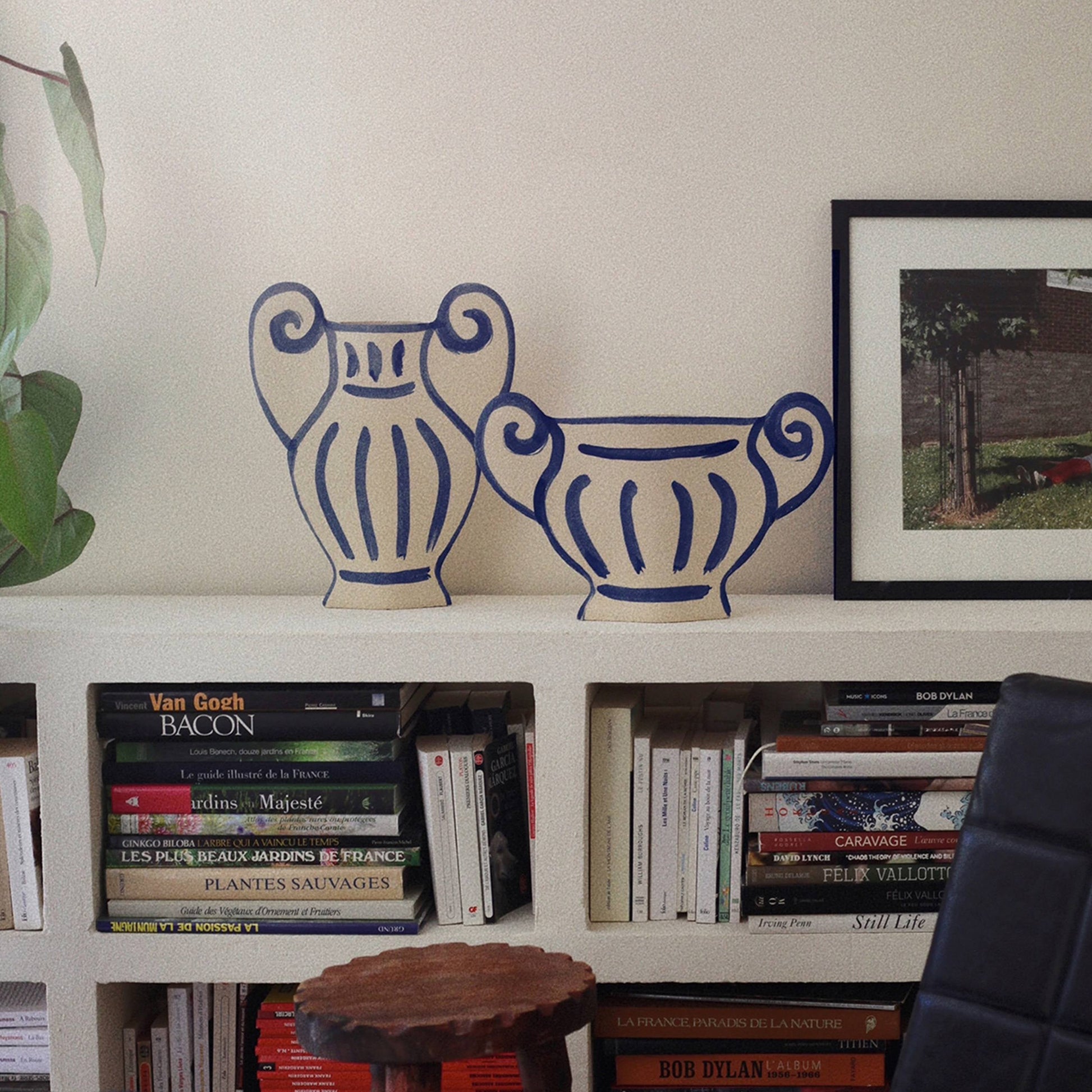 Two INI CERAMIQUE Greek Coupe Ceramic Vases with decorative handles rest on a white shelf beside a framed photo, surrounded by art books and a green plant, bringing timeless Greek-inspired elegance to your decor.
