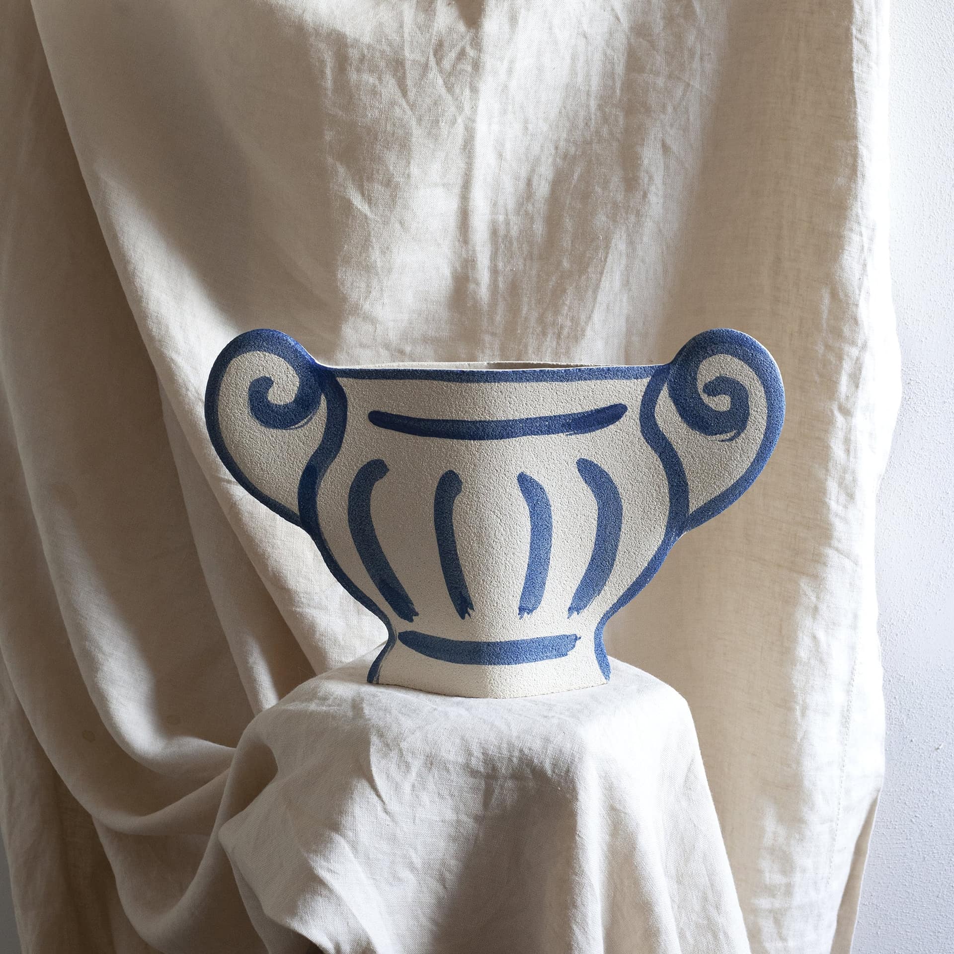 The INI CERAMIQUE Greek Coupe Ceramic Vase by INI CERAMIQUE features a white, handcrafted design with blue lines and two large curved handles, evoking ancient Greek pottery and styled like a textured trophy or urn. Displayed against light-colored fabric.