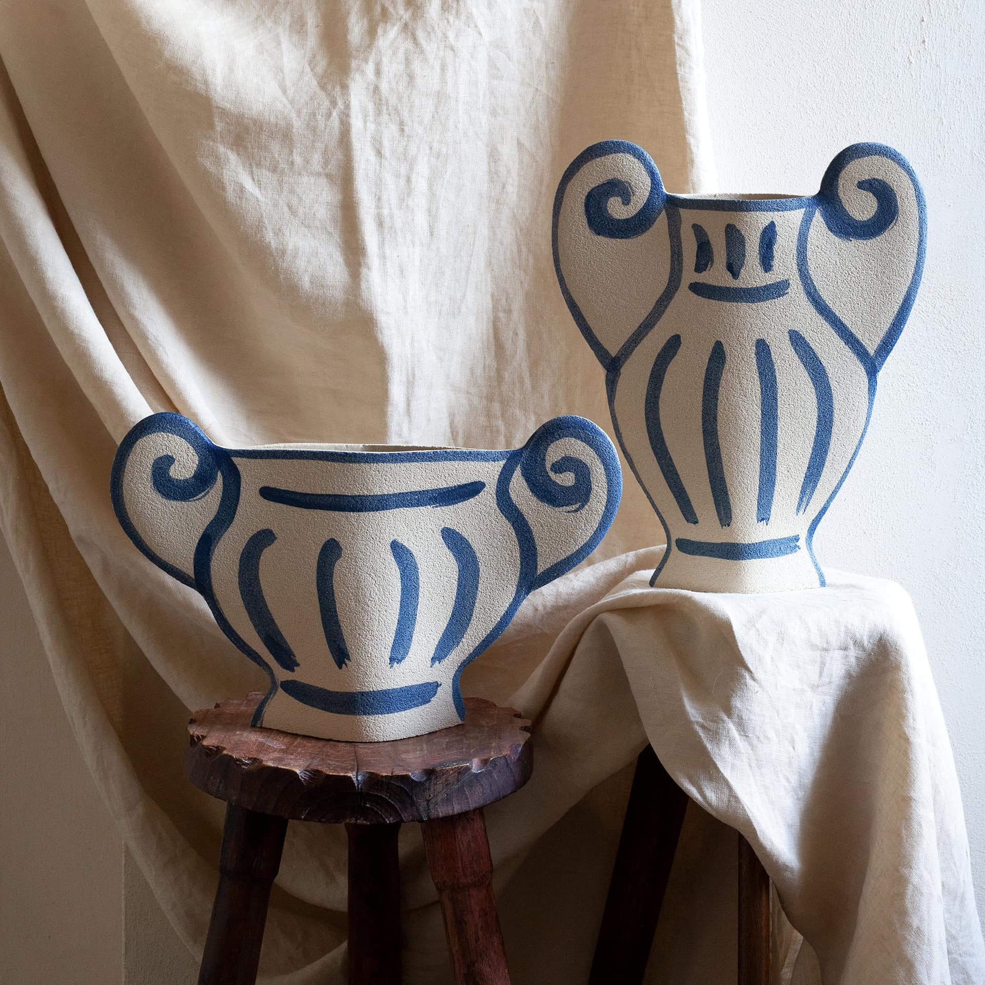 Two INI CERAMIQUE Greek Coupe Ceramic Vases with blue, stylized designs and curved handles sit on wooden stools against a beige fabric backdrop, evoking the look of ancient Greek pottery.