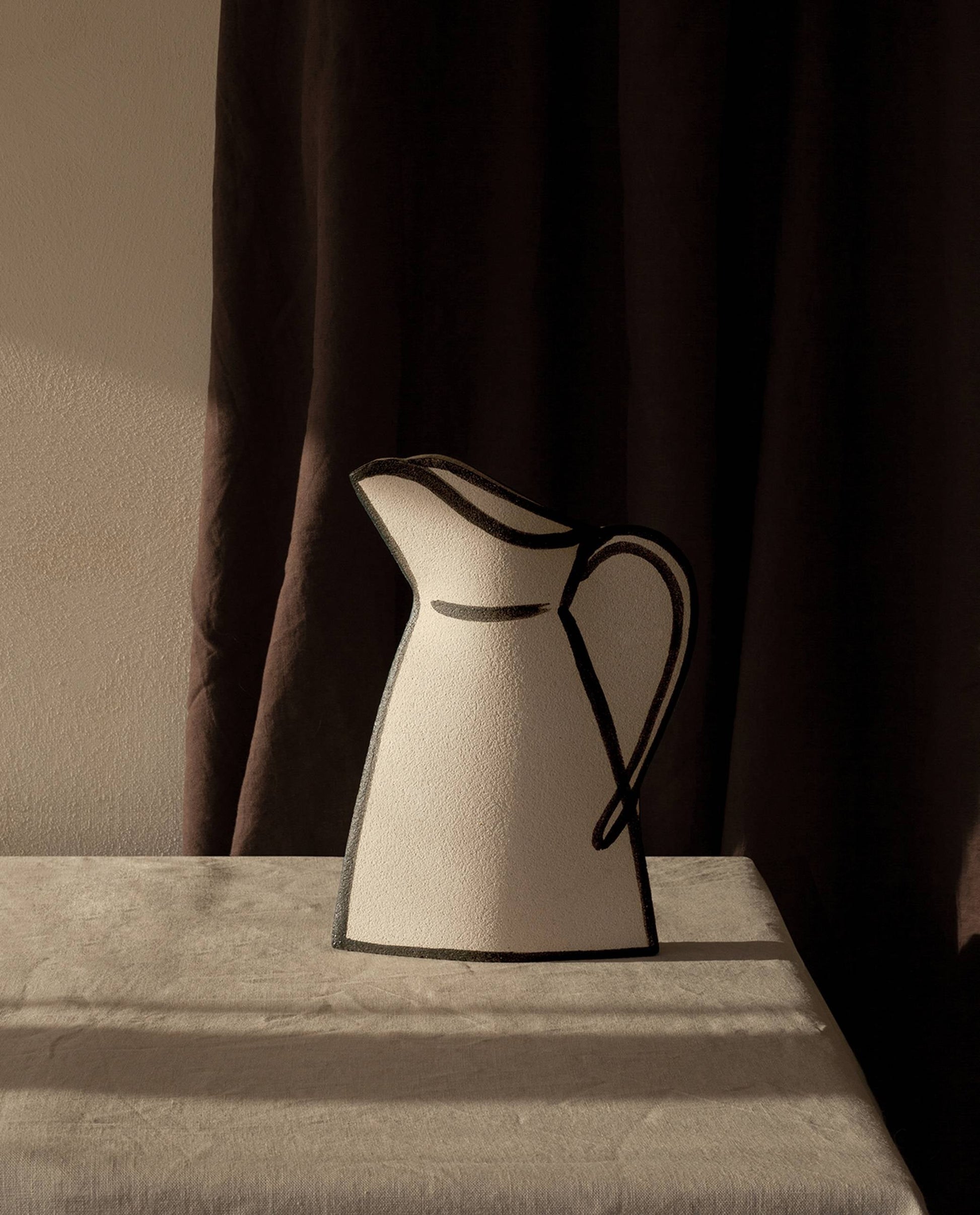 The INI CERAMIQUE Morandi Pitcher Ceramic Vase with black outlines rests on a beige cloth-covered table, its artisanal craftsmanship accentuated by dramatic light and shadows against a softly lit wall and dark curtain backdrop.