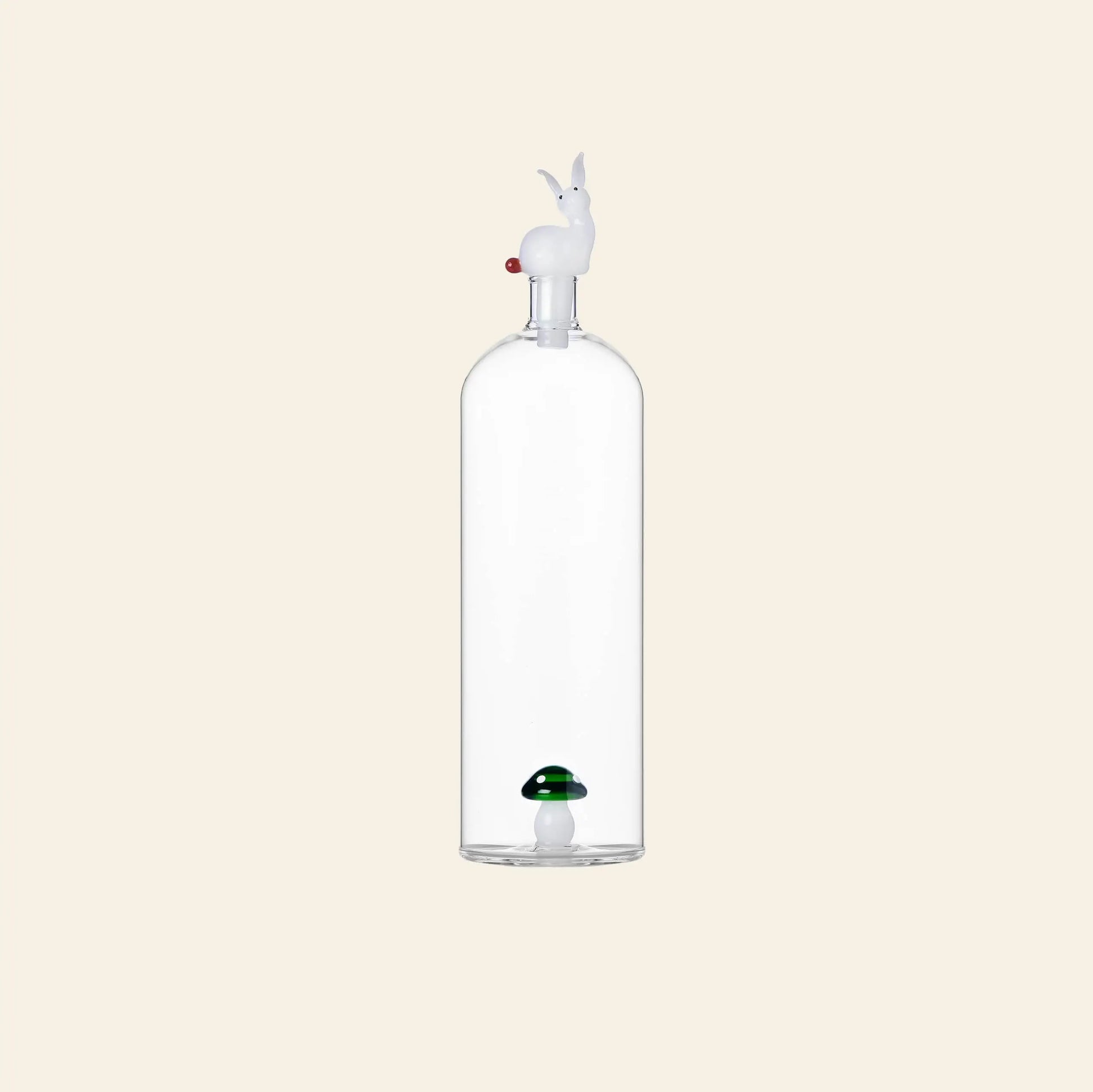 The Ichendorf Milano Alice Bottle by Ichendorf Milano features clear glass, a white rabbit-shaped stopper, and a small green and white mushroom figurine inside, creating whimsical wonderland charm against a plain beige background.