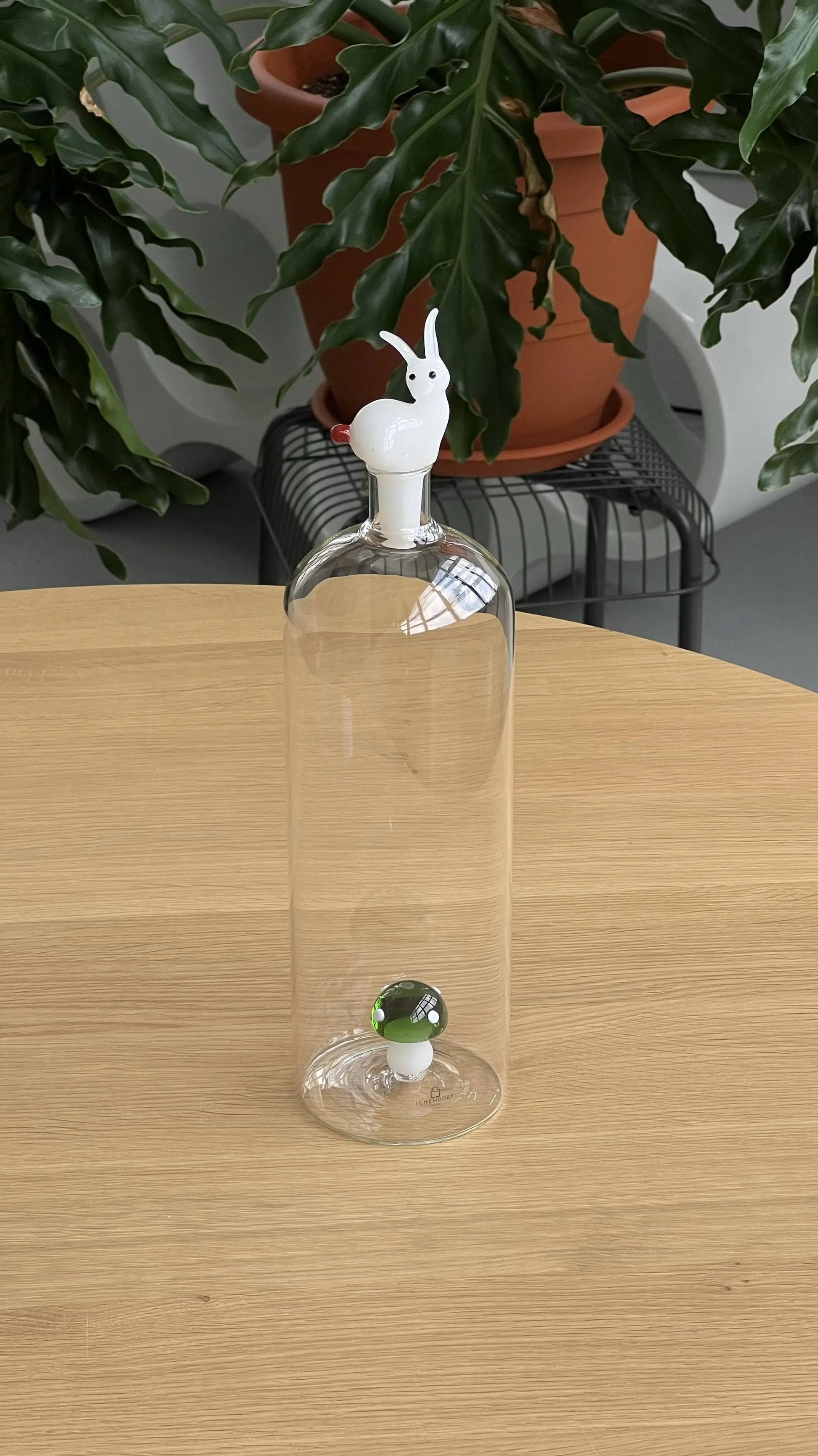 The Ichendorf Milano Alice Bottle by Ichendorf Milano features a clear glass design with a white animal-shaped stopper, possibly a rabbit. It sits on a light wood table with a green and white sphere inside, while potted plants create a whimsical atmosphere.