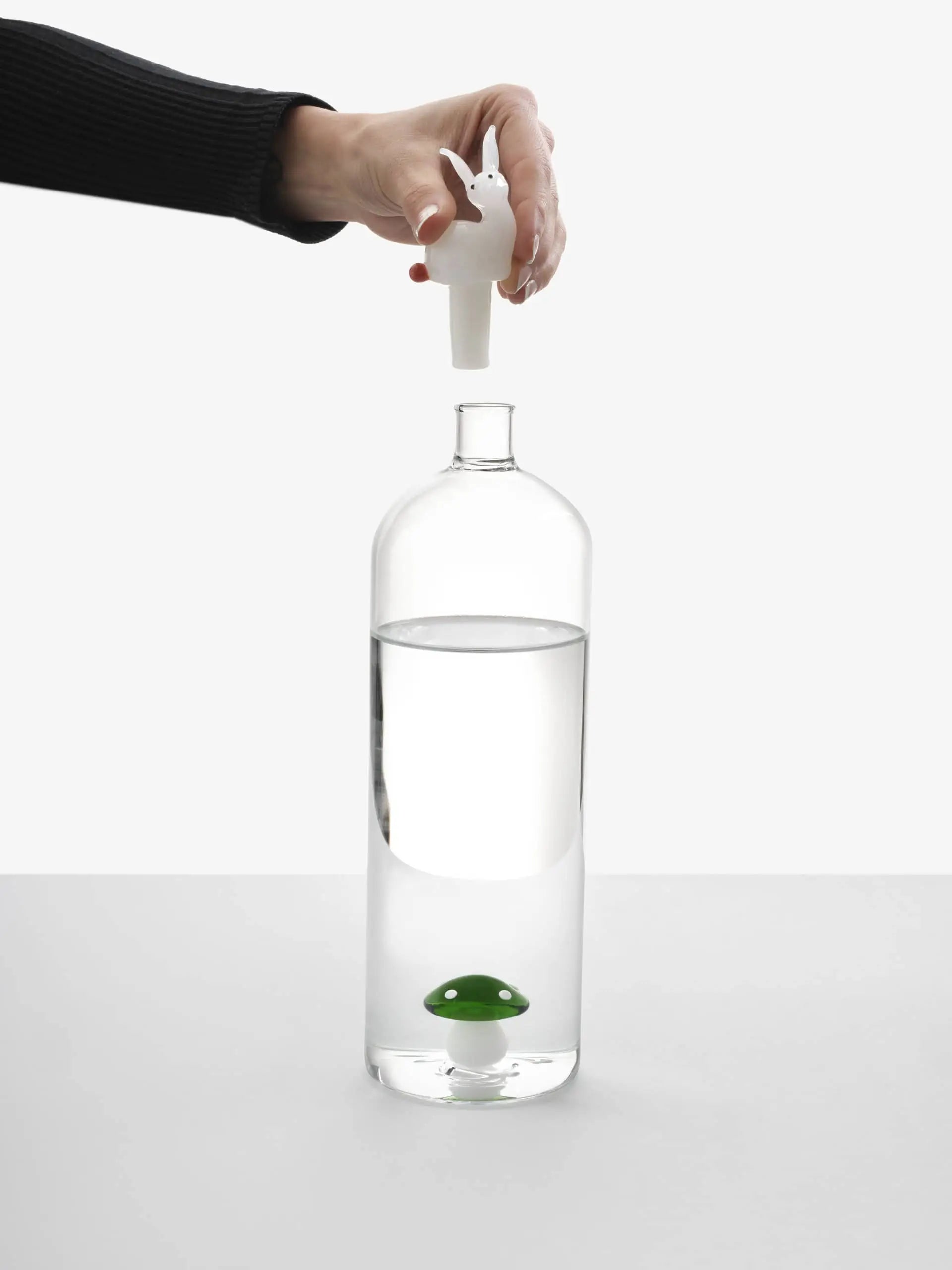 A hand holds a white rabbit-shaped stopper above the Ichendorf Milano Alice Bottle by Ichendorf Milano, which is filled with water and features a floating green frog inside, adding a whimsical charm to the unique glassware.