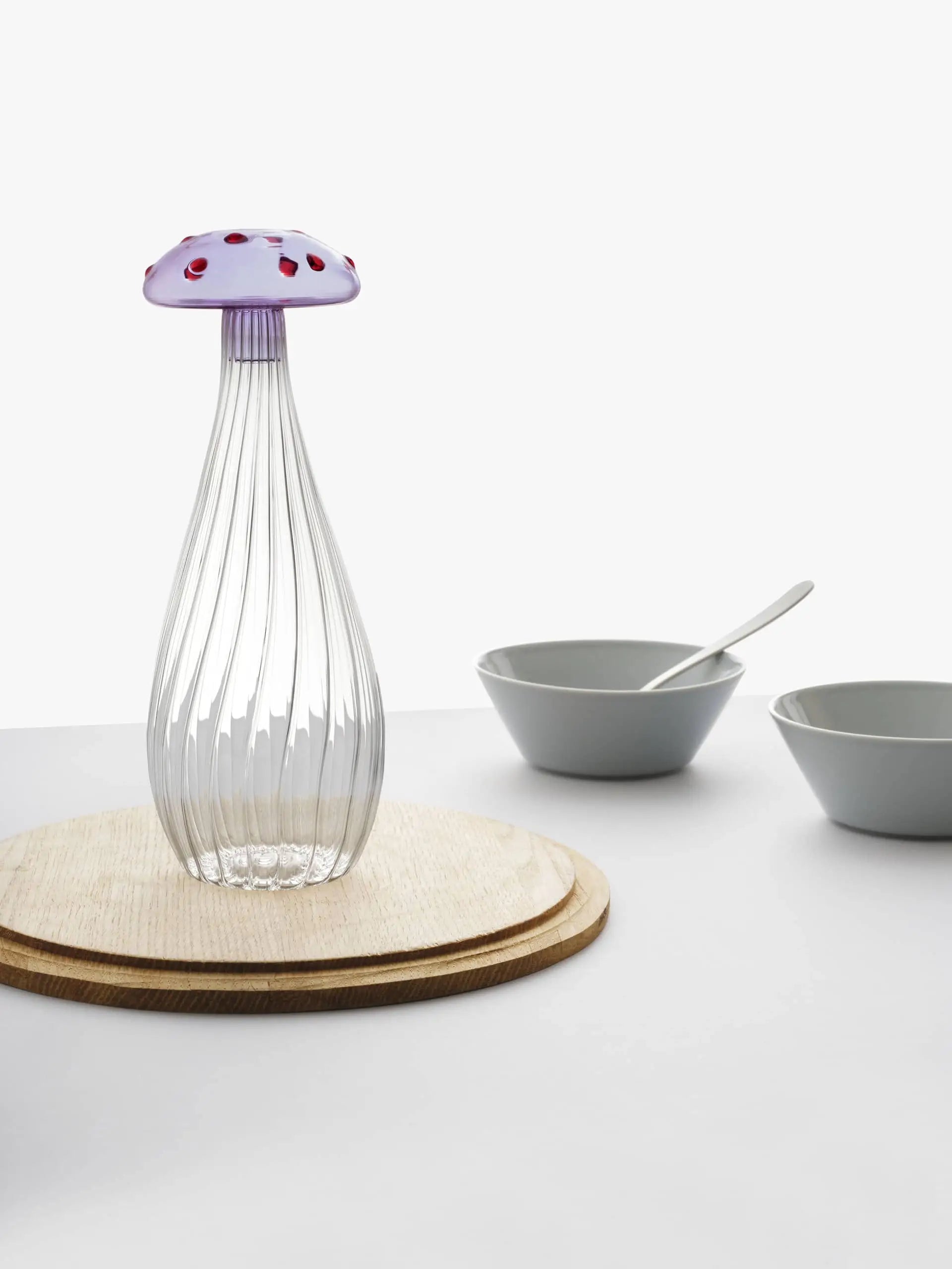 The Ichendorf Milano Alice Bottle, shaped like a colorful mushroom with a purple and red-spotted top and clear ribbed stem, sits on round wooden boards. Two white bowls and a spoon rest nearby against a plain background.