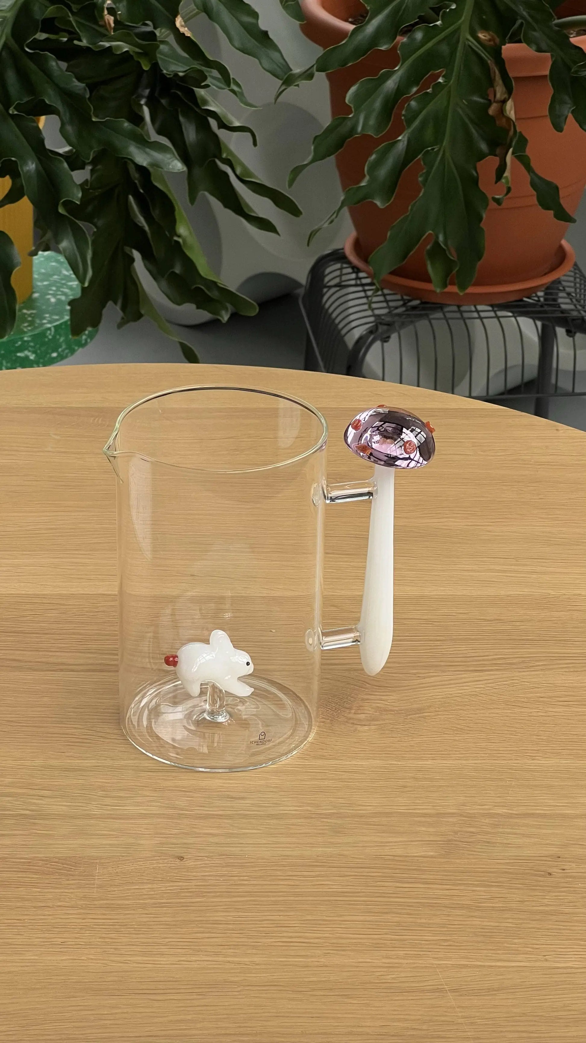 The Ichendorf Milano Alice Jug is a clear, handcrafted glass jug featuring a handle shaped like a white mushroom with a pink and purple cap. Inside sits a small white glass dog, while green plants enhance its whimsical wonderland feel.