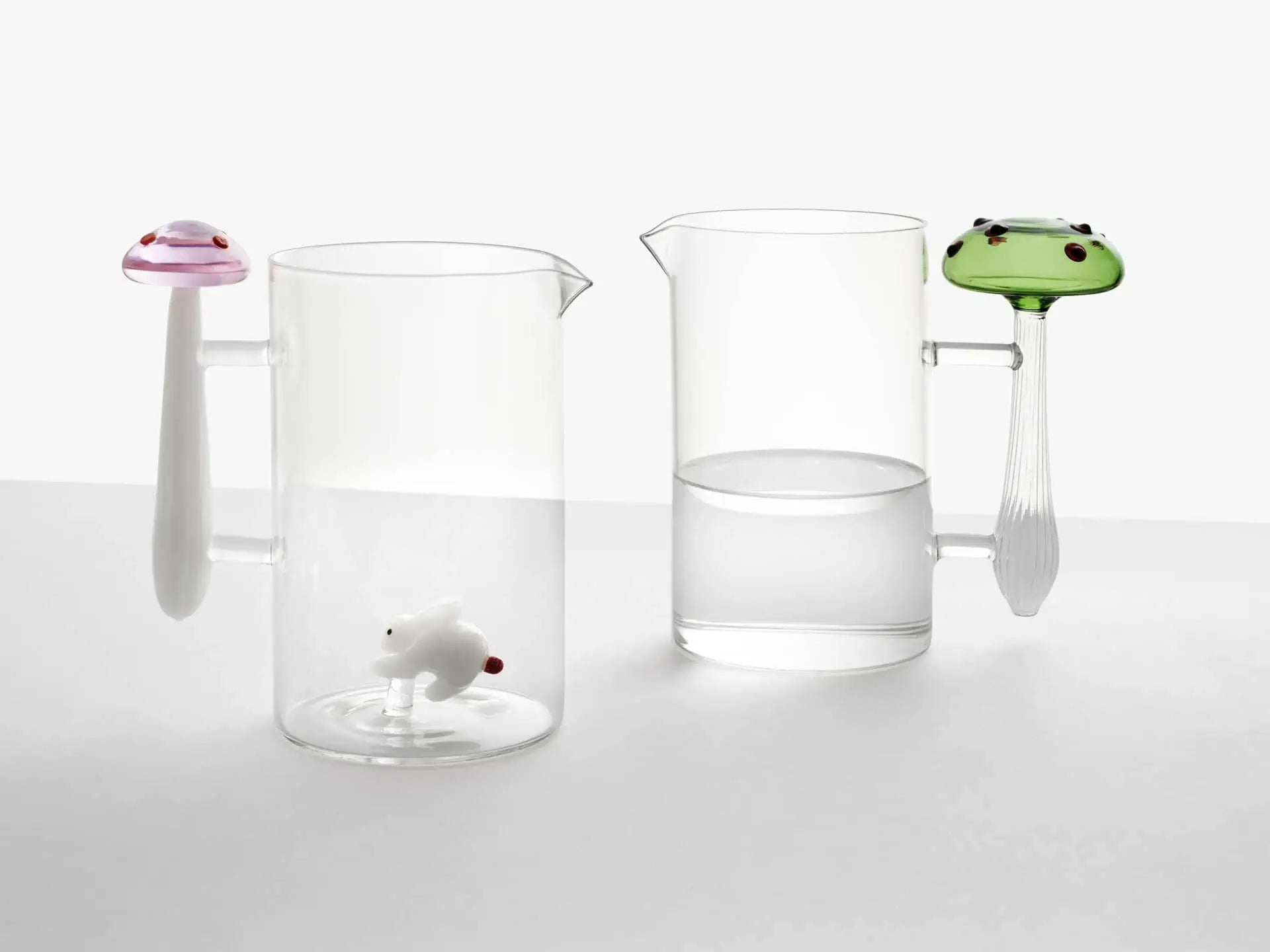 Two Ichendorf Milano Alice Jugs sit side by side; one has a pink mushroom-shaped handle with a tiny white animal inside, and the other features a green frog-shaped handle and is half-filled with water.