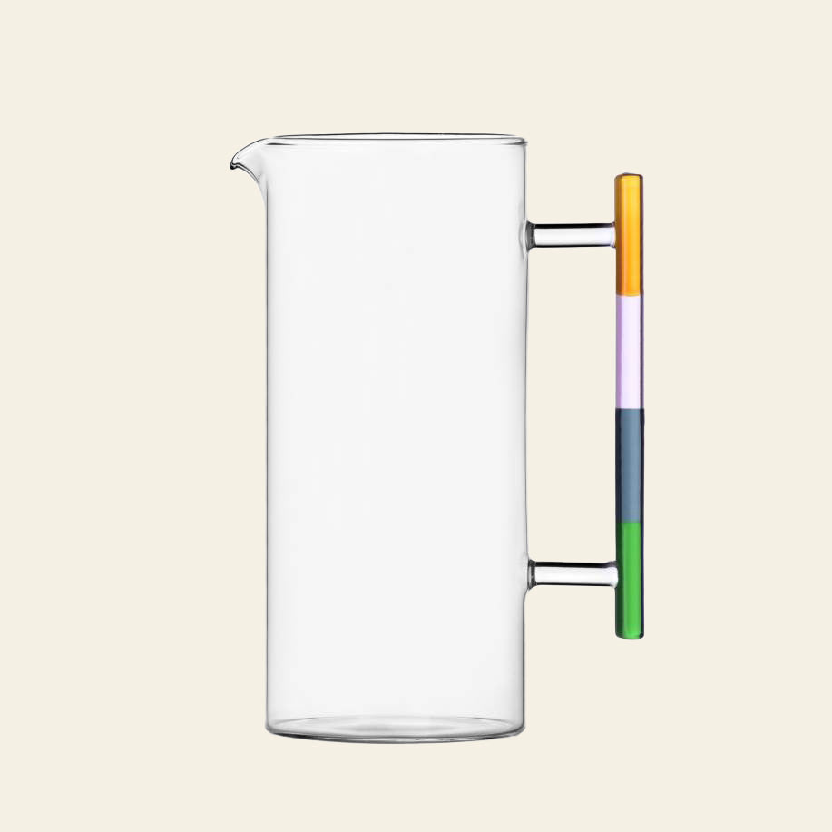 The Ichendorf Milano Andalusia Jug is a clear, handmade borosilicate glass pitcher with a cylindrical shape, straight spout, and a colorblocked gradient handle in stacked yellow, orange, purple, blue, and green.