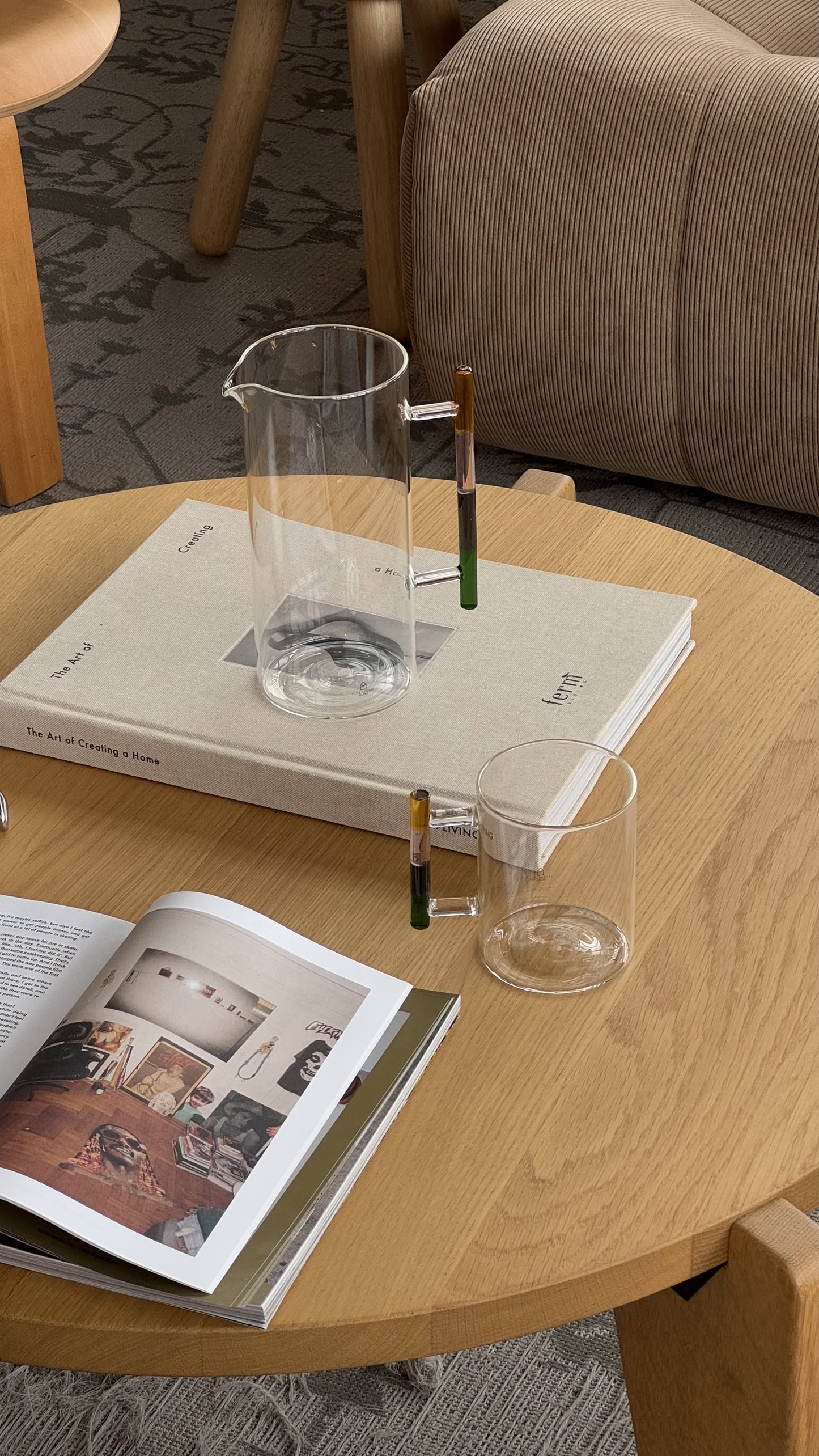 An Ichendorf Milano Andalusia Jug with a matching cup, both featuring colorblocked gradient handles, sit on a light wood coffee table beside books in a cozy living room with neutral furniture and a patterned carpet.