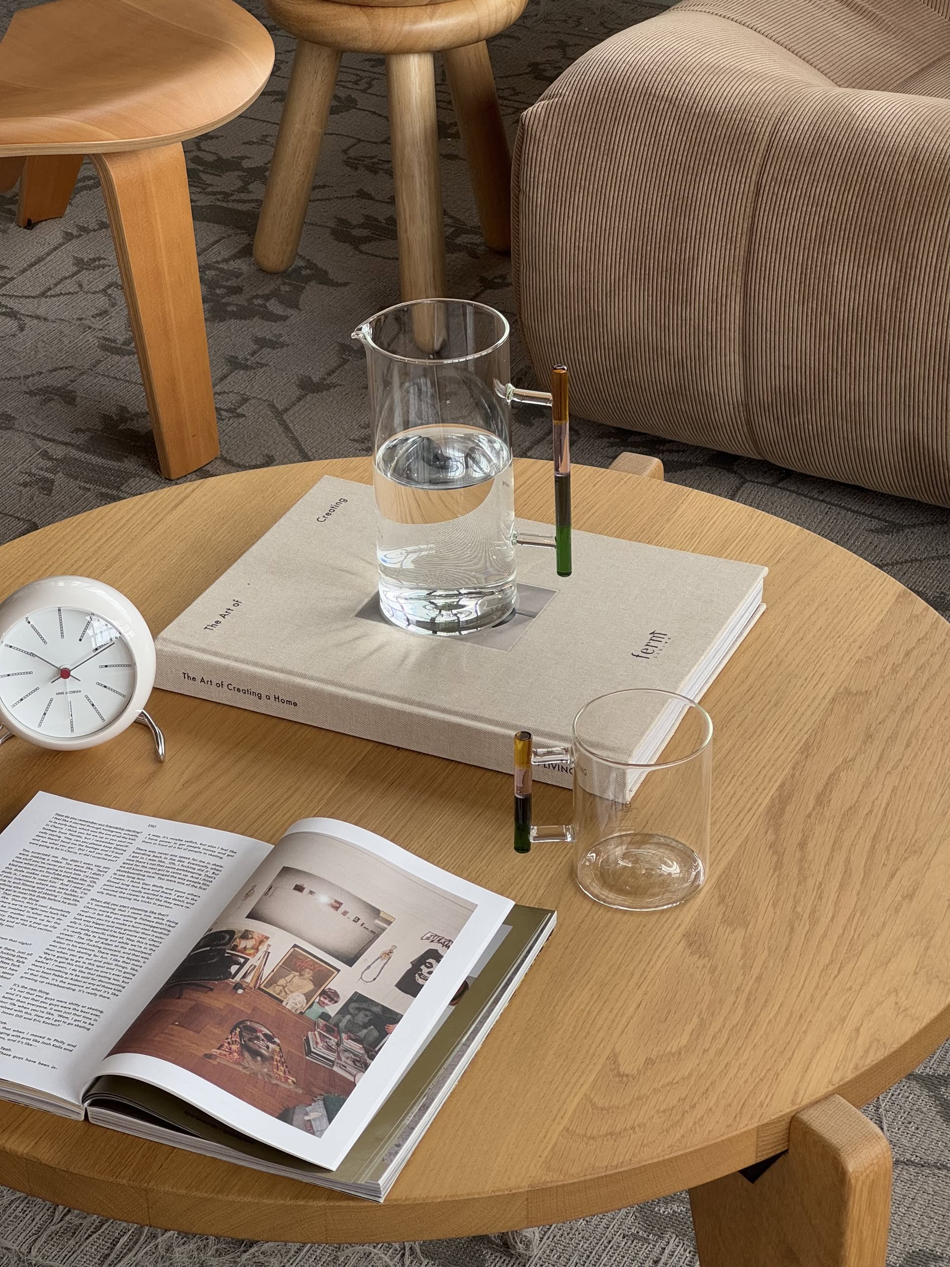 A modern wooden coffee table displays the Ichendorf Milano Andalusia Jug, a handmade borosilicate glass cup, a book, an open magazine, and a white clock. In the background are a beige armchair and wooden stools on a patterned carpet.