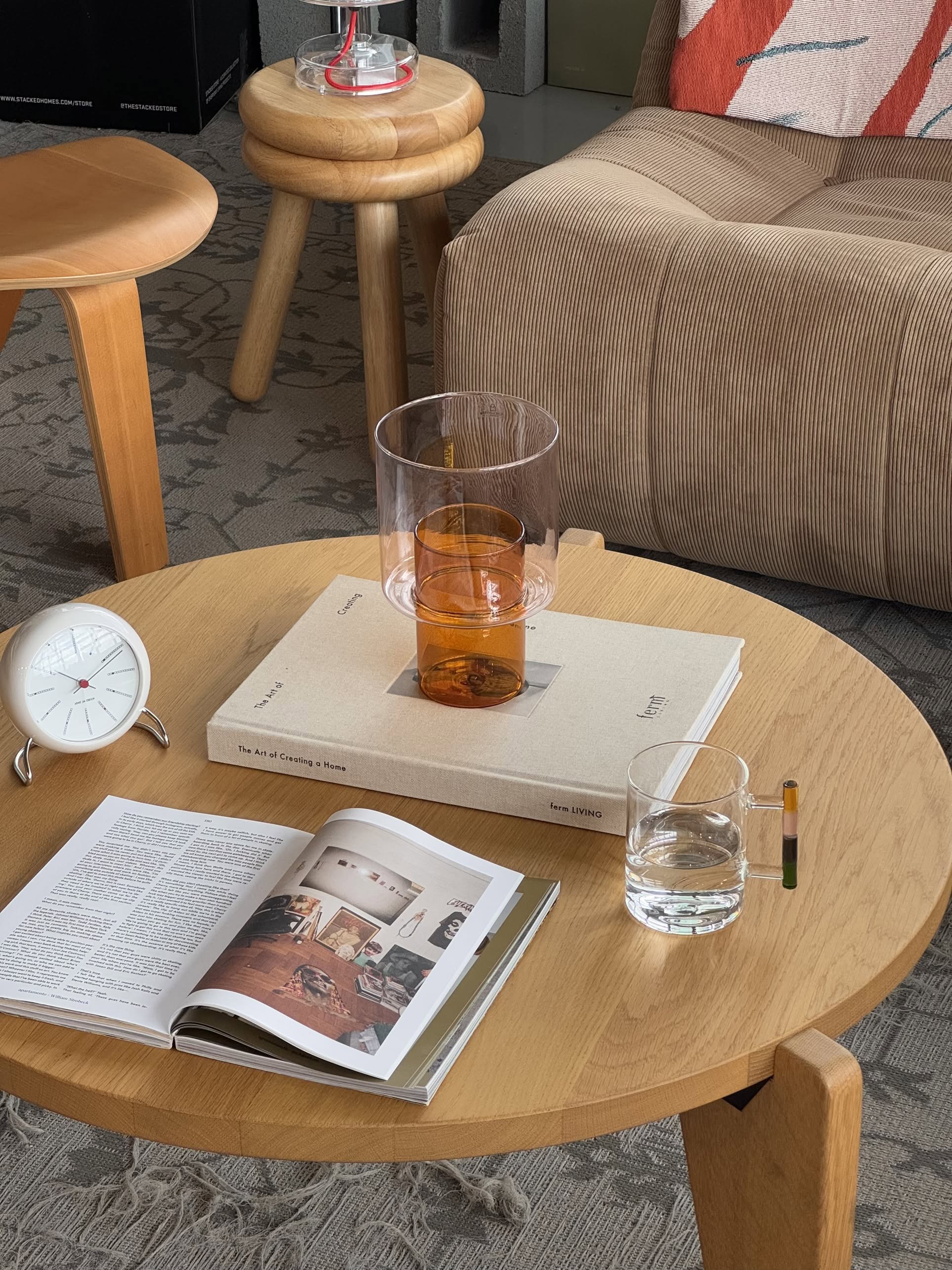 A cozy coffee table holds an open art book, a glass of water, two colored pencils, and the Ichendorf Milano Andalusia Mug by Ichendorf Milano with a colourblocked handle atop a closed hardcover book. Modern chairs and a textured rug are in the background.