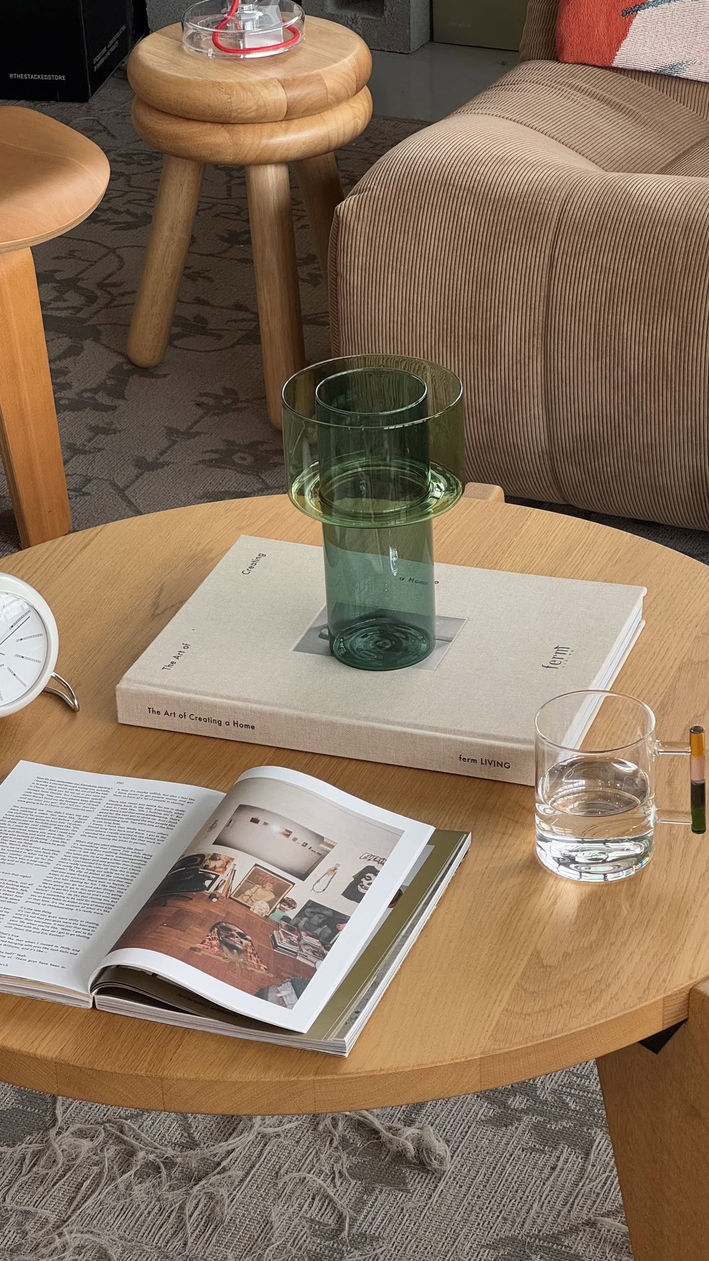 A round wooden coffee table with a glass of water, books, a green glass vase, a pencil, and the Ichendorf Milano Andalusia Mug by Ichendorf Milano sits in a cozy living room featuring a patterned rug and cushioned seating.