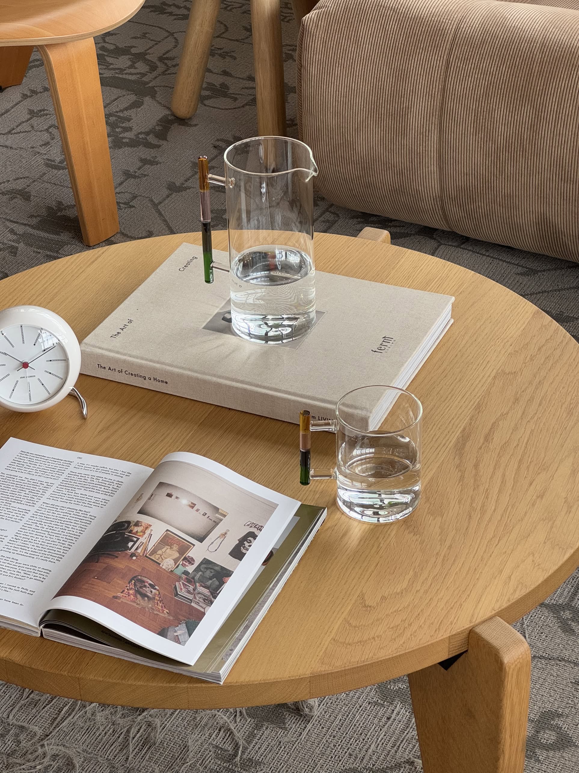 A round wooden coffee table holds an open magazine, a closed book, two pens, a small white clock, and an Ichendorf Milano Andalusia Mug by Ichendorf Milano. Upholstered furniture and a patterned rug are seen in the background.