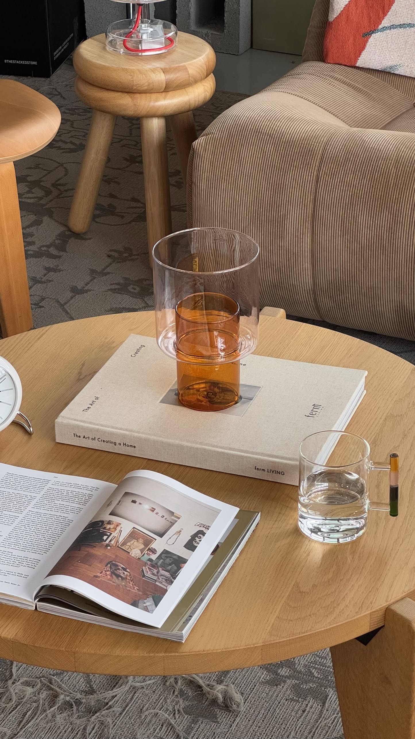 A wooden coffee table holds a closed book, an open magazine, and the Ichendorf Milano Andalusia Mug by Ichendorf Milano stacked inside a clear glass of water. Cozy furniture and a textured rug complete the inviting scene.