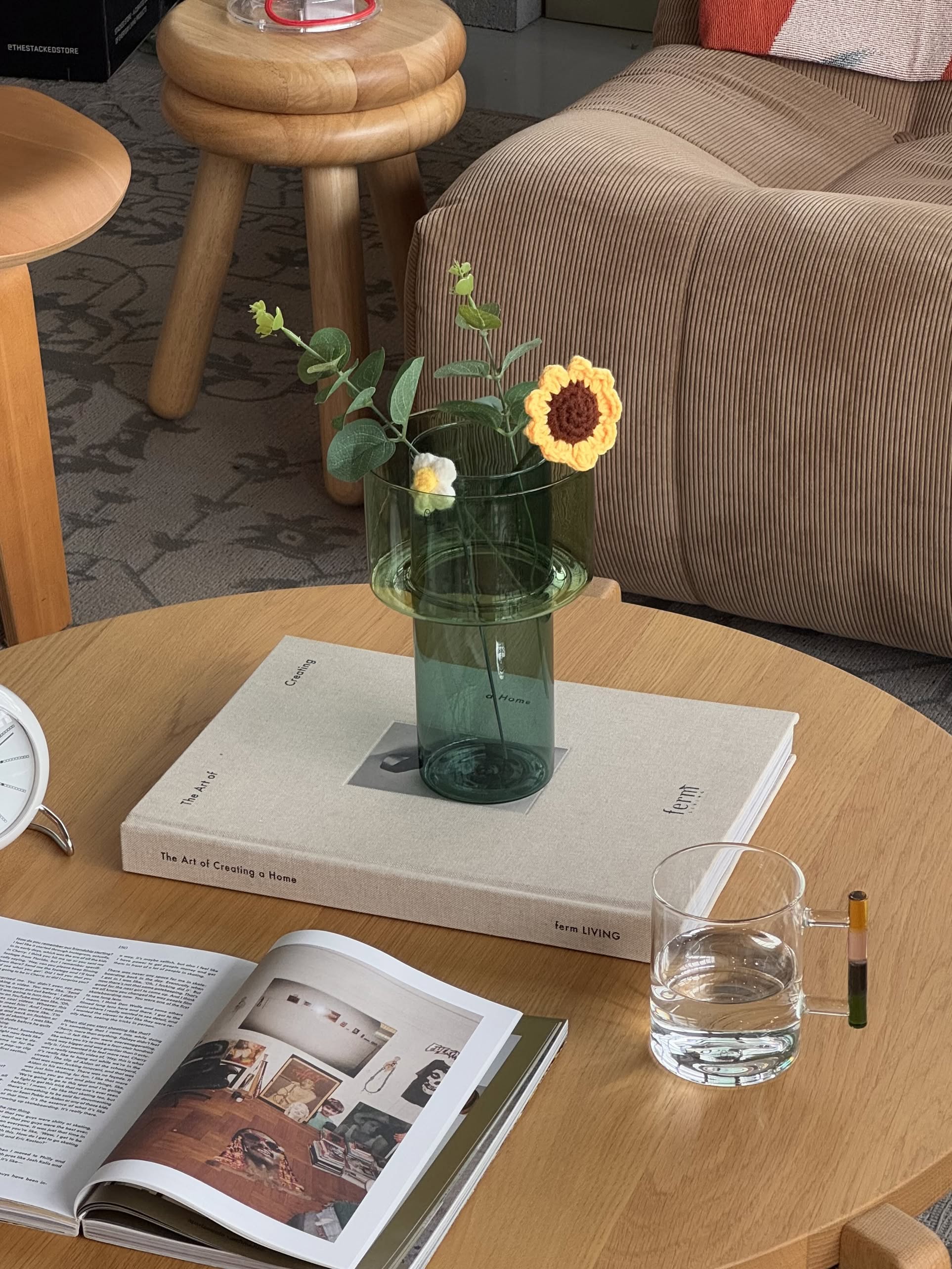 A green glass vase with crocheted flowers sits on a beige book atop a wooden table; nearby are an open magazine, yellow pen, and Ichendorf Milano Andalusia Mug, featuring a colourblocked handle and handmade borosilicate glass by Ichendorf Milano.